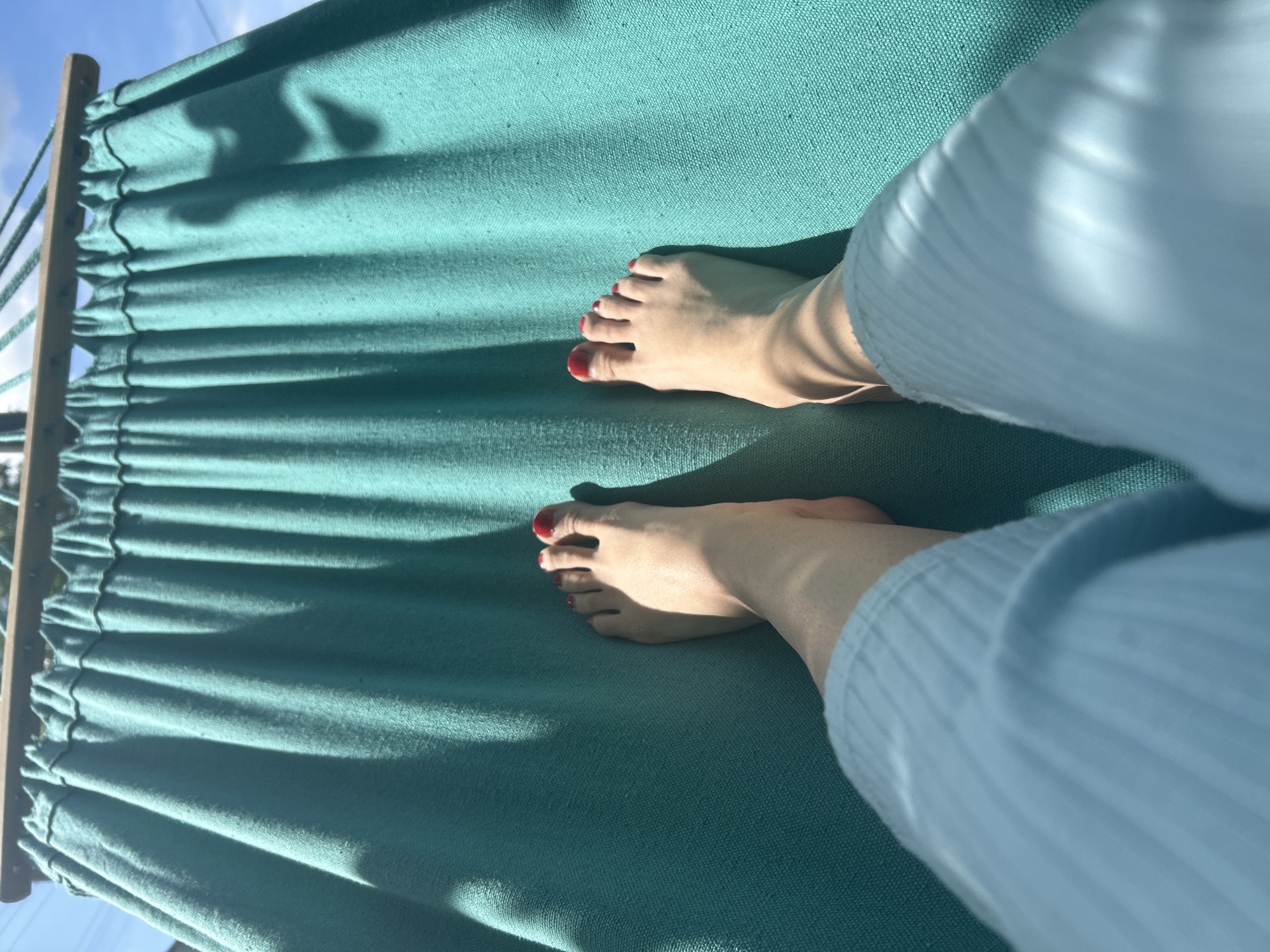 A first-person perspective shot captures two bare feet, each with red nail polish, resting on a teal-colored fabric hammock. The hammock is suspended by a wooden bar and ropes, and sunlight casts dappled shadows across its surface. The person appears to be wearing light blue, ribbed pajama pants. The background consists of a clear blue sky and hints of greenery, suggesting an outdoor setting. The image was taken in Vyshevychi, Ukraine.