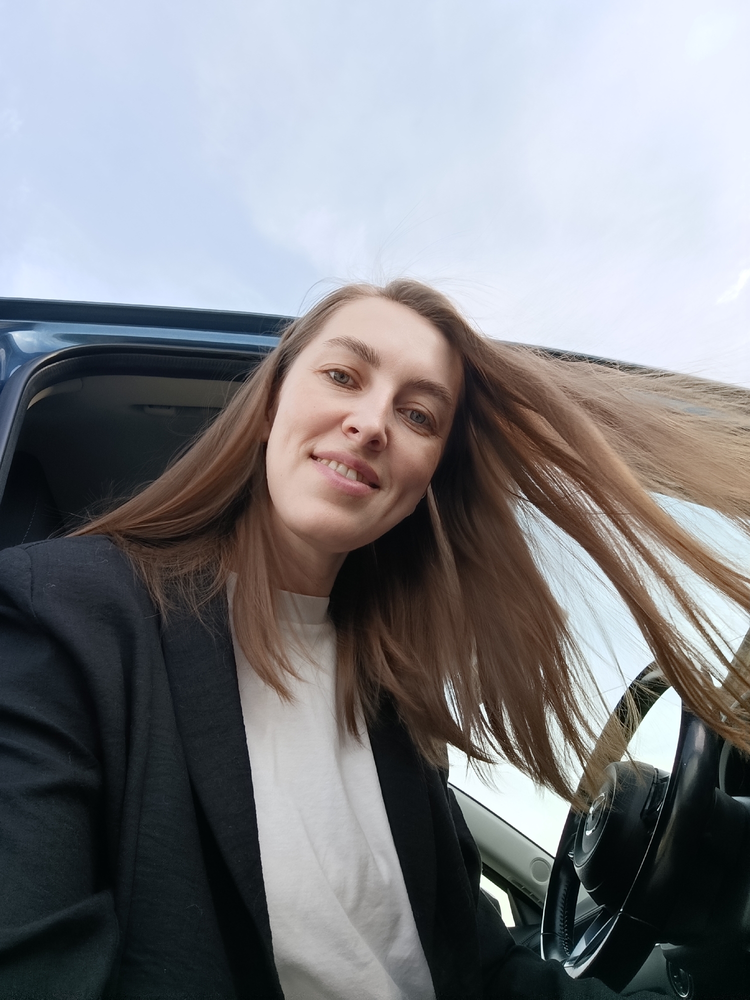 A woman is seated inside a vehicle, possibly the driver's seat, with the steering wheel visible to the right. She is wearing a black blazer over a white t-shirt. Her hair is long and brown, blowing to one side. The sky is visible through the car window, suggesting an outdoor setting. No specific details indicate the city of Chaikovsky, Russia, are present in the image.