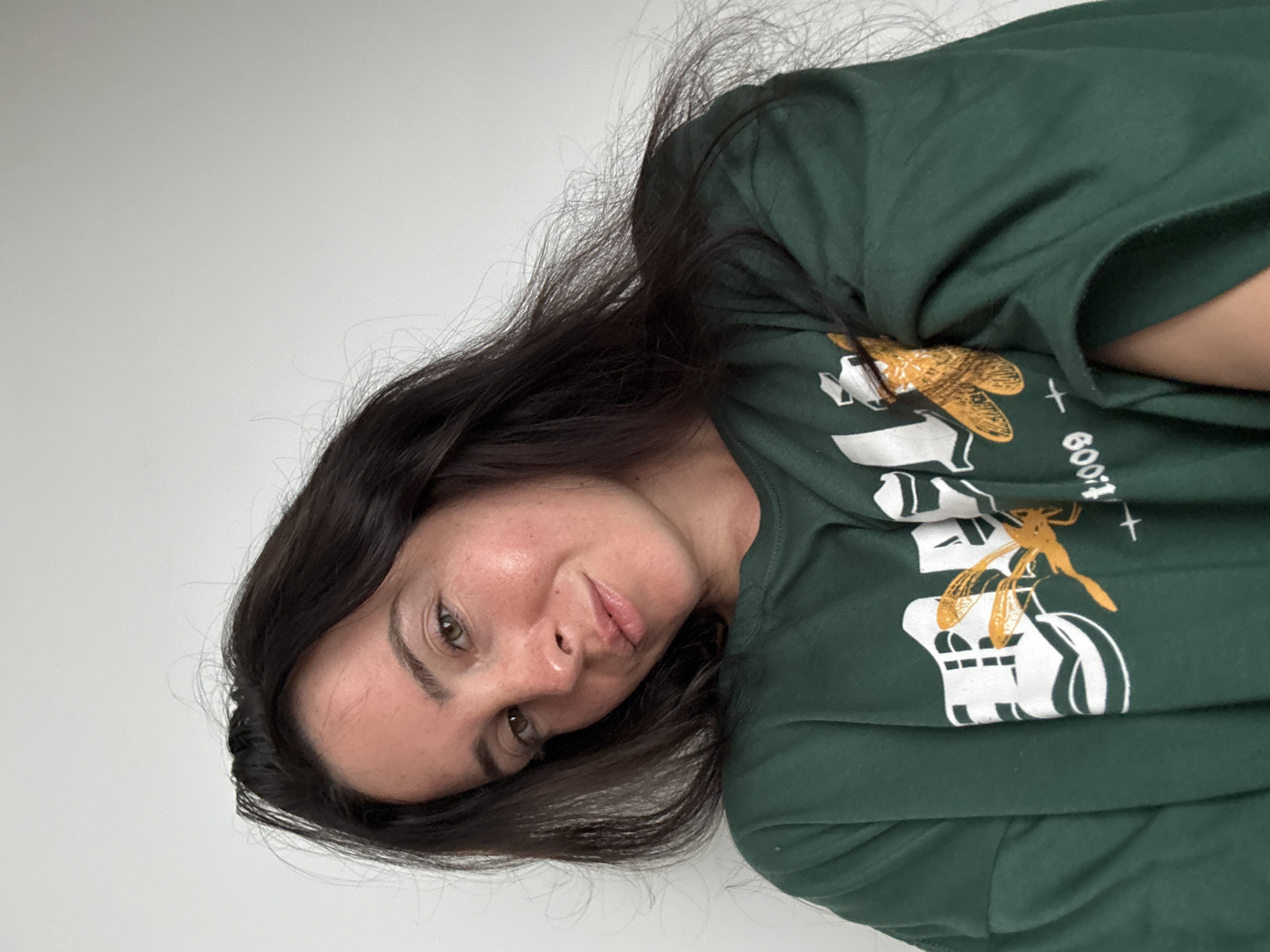 A young woman with long, dark brown hair, wearing a green t-shirt with a graphic print, poses for a selfie against a white wall. The graphic on her shirt features white lettering and a depiction of a dragonfly. She has fair skin with some redness on her cheeks and nose, and her eyes are green. She is looking at the camera with a slight smile. The background is plain and uncluttered, suggesting an indoor setting. There is no visible indication of the city of North Kuta, Indonesia.