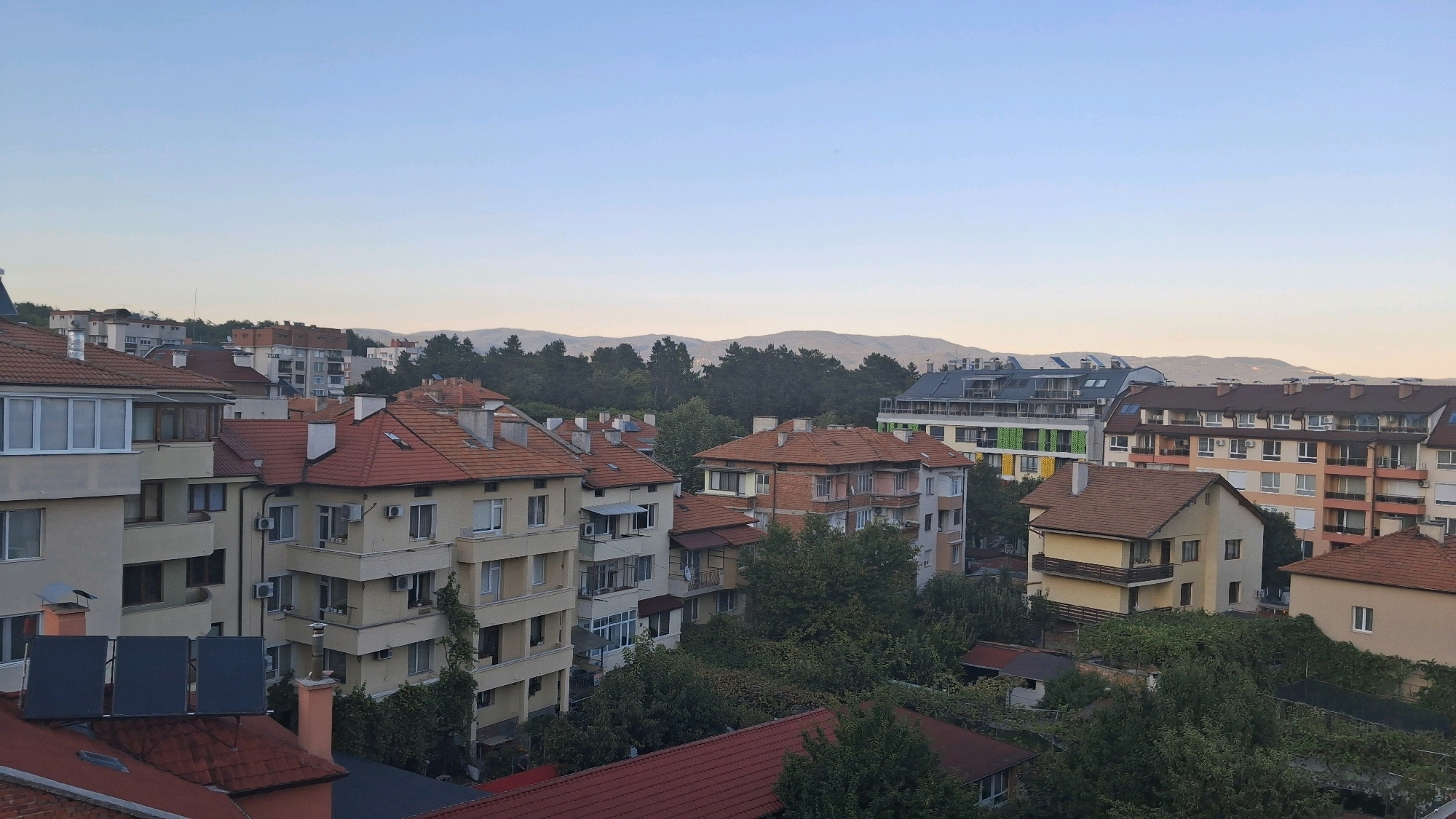 This is a wide, high-angle shot of a residential area in Gotse Delchev, Bulgaria, taken at dawn. The sky is a pale blue, transitioning to a lighter hue near the horizon, indicating the early morning light. In the foreground, several buildings with terracotta tiled roofs are visible. These are multi-story apartment buildings with a mix of cream and yellow facades, some with balconies and air conditioning units. Interspersed among the buildings are numerous mature trees, some with lush green foliage, others appearing more sparse. In the middle ground, more apartment buildings can be seen, including one with a distinctive green, white, and yellow facade. Further in the background, a dense line of dark green coniferous trees forms a backdrop, beyond which lie rolling hills and mountains, softened by the morning mist. The overall impression is one of a quiet, established urban neighborhood on the edge of a natural landscape, bathed in the soft light of sunrise.