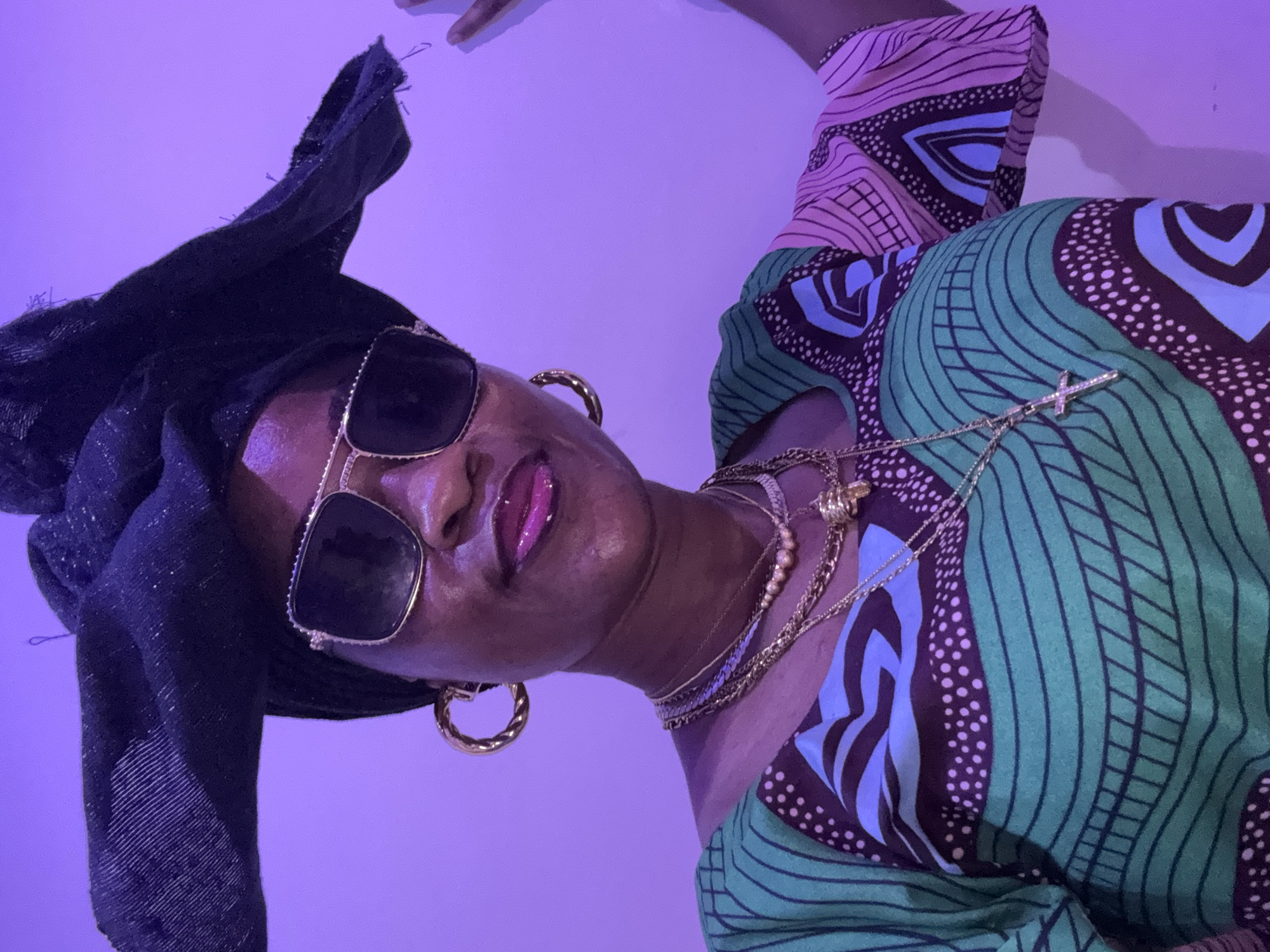 The image shows a close-up, vertically oriented, of a person wearing a black headscarf, oversized sunglasses, and a vibrantly patterned green and pink outfit. The person has on multiple gold necklaces, including one with a cross pendant, and large gold hoop earrings. Their lips are glossy and appear to be painted with pink lipstick. The background is a solid, light purple color. The lighting suggests an indoor setting, possibly illuminated by artificial lights, with a purplish hue cast over the scene. There are no visible signs of text or specific location cues within the frame, though the prompt provides "Ajara, Nigeria" as context. The overall mood conveyed is one of confidence and style, with a focus on fashion accessories and the colorful attire. The person's pose and expression suggest they are posing for a photograph.