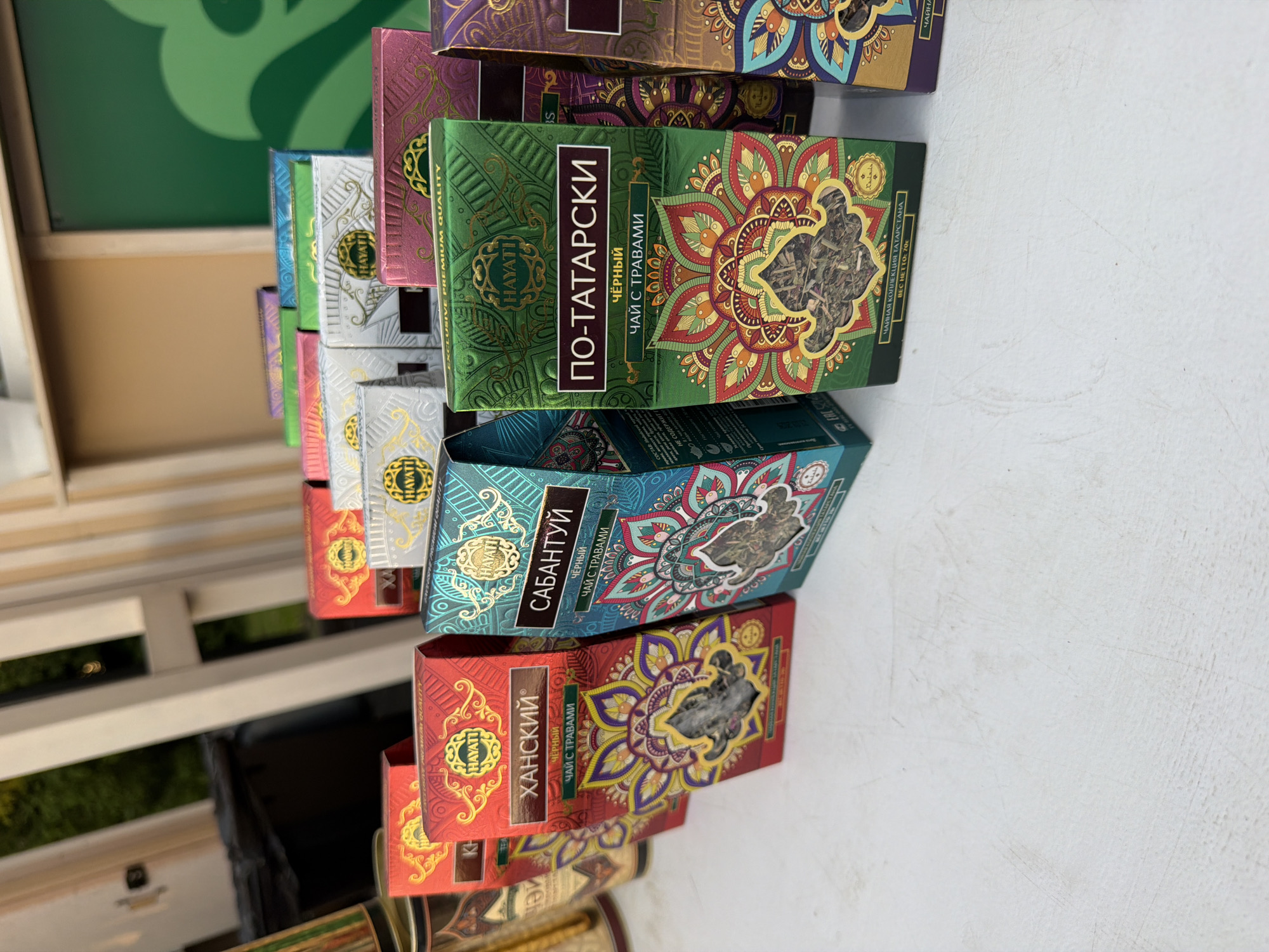 The image shows a close-up, eye-level shot of a collection of tea boxes arranged on a white, textured surface. The boxes are colorful and feature intricate, mandala-like designs with gold accents. Some of the boxes are open, revealing the tea leaves inside. The tea boxes are labeled in Russian, with prominent branding for "HAYATI". The lighting is natural and suggests an outdoor setting, possibly a market or a display in a shop, given the blurred background of wooden structures and greenery. The perspective is slightly angled, with the boxes receding into the background.