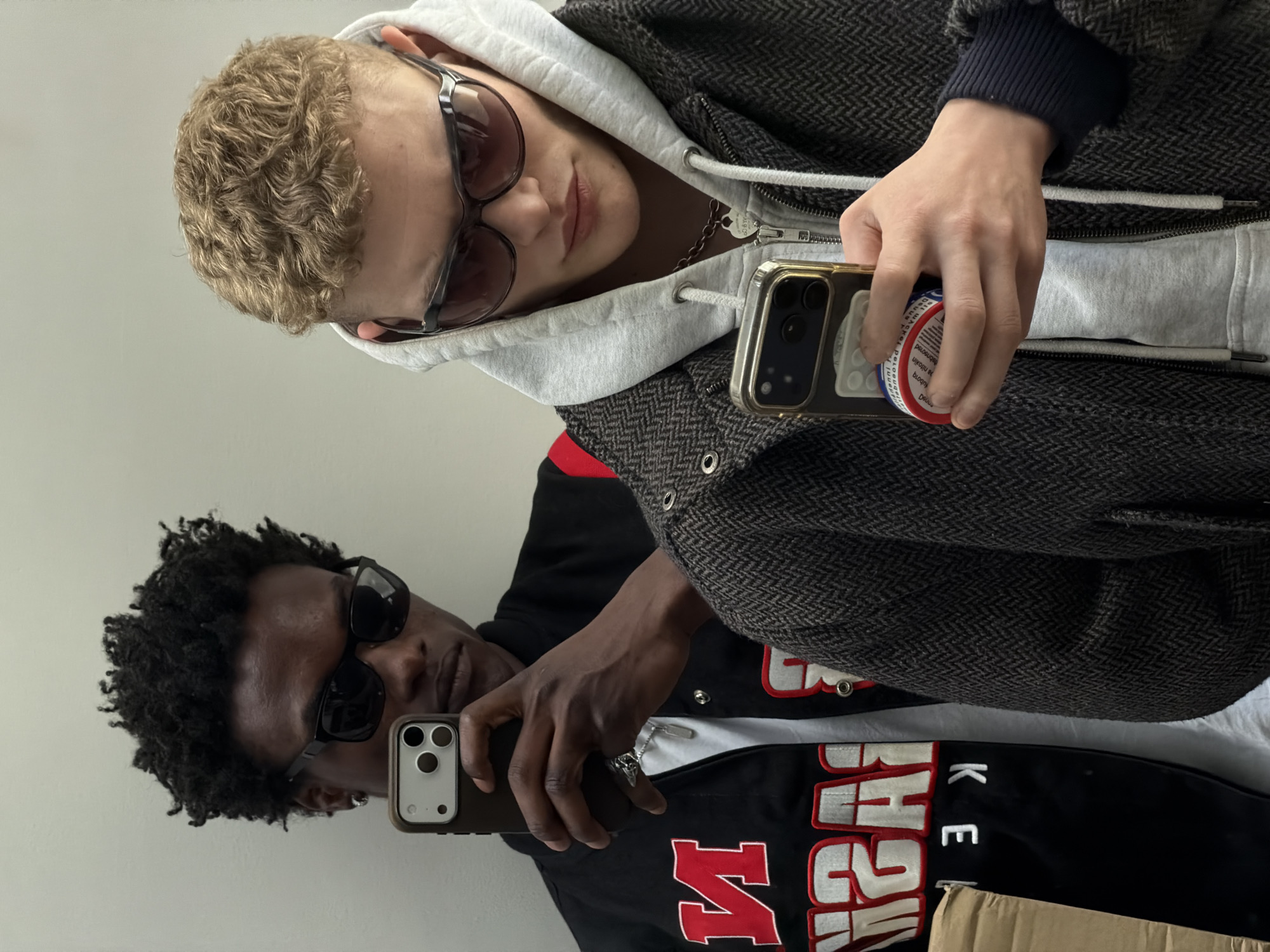 Two individuals are taking a selfie in what appears to be an indoor setting.
The first individual, fair-skinned with short, curly blonde hair, is wearing sunglasses, a grey hooded sweatshirt, and a patterned jacket. They are holding a phone with a pop socket.
The second individual is dark-skinned with curly black hair and is also wearing sunglasses. They are holding a different phone. They are wearing a black jacket with red and white lettering, partially obscured.
The scene lacks distinctive architectural features or signage to identify a specific location in London. The background is a plain white wall.
