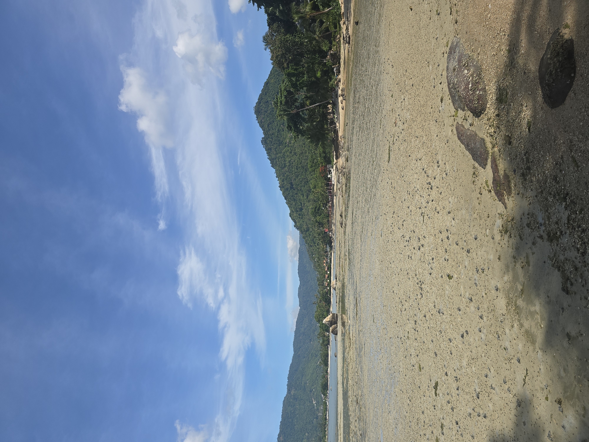 The image depicts a low tide scene at a beach in Baan Chaweng Noi, Thailand. The foreground is dominated by wet, sandy ground dotted with small pebbles and occasional larger rocks, some of which are submerged in shallow water. In the middle ground, more rocks and some patches of darker sand are visible, with a body of calm water extending towards the background. The background features a lush, green, tree-covered hillside that slopes down to the coast. A few buildings and palm trees are sparsely visible along the shoreline, suggesting human habitation. The sky above is a clear blue with scattered white clouds. The overall scene is tranquil and natural, with no people or distinct actions depicted.