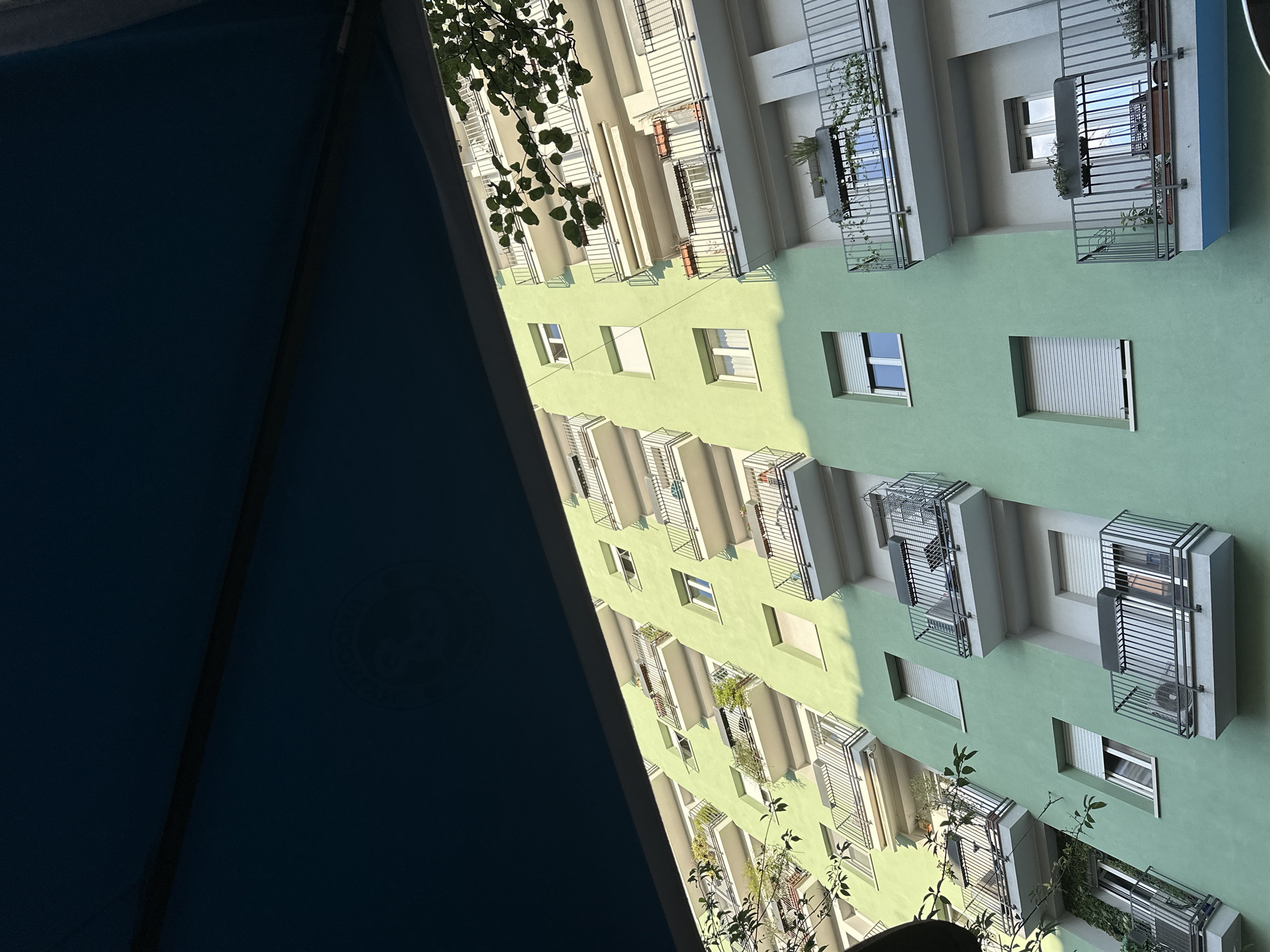 The image shows an upward-angled view of a building facade in Milan, Italy. The building has a light green exterior with multiple windows, some of which are fitted with metal cages or balconies. Plants are visible on some balconies. The photograph is taken from a low vantage point, possibly from a courtyard or balcony. A dark blue, tent-like structure occupies a significant portion of the left side of the frame, partially obscuring the view. The lighting suggests it is daytime, with sunlight casting shadows on the building facade.
