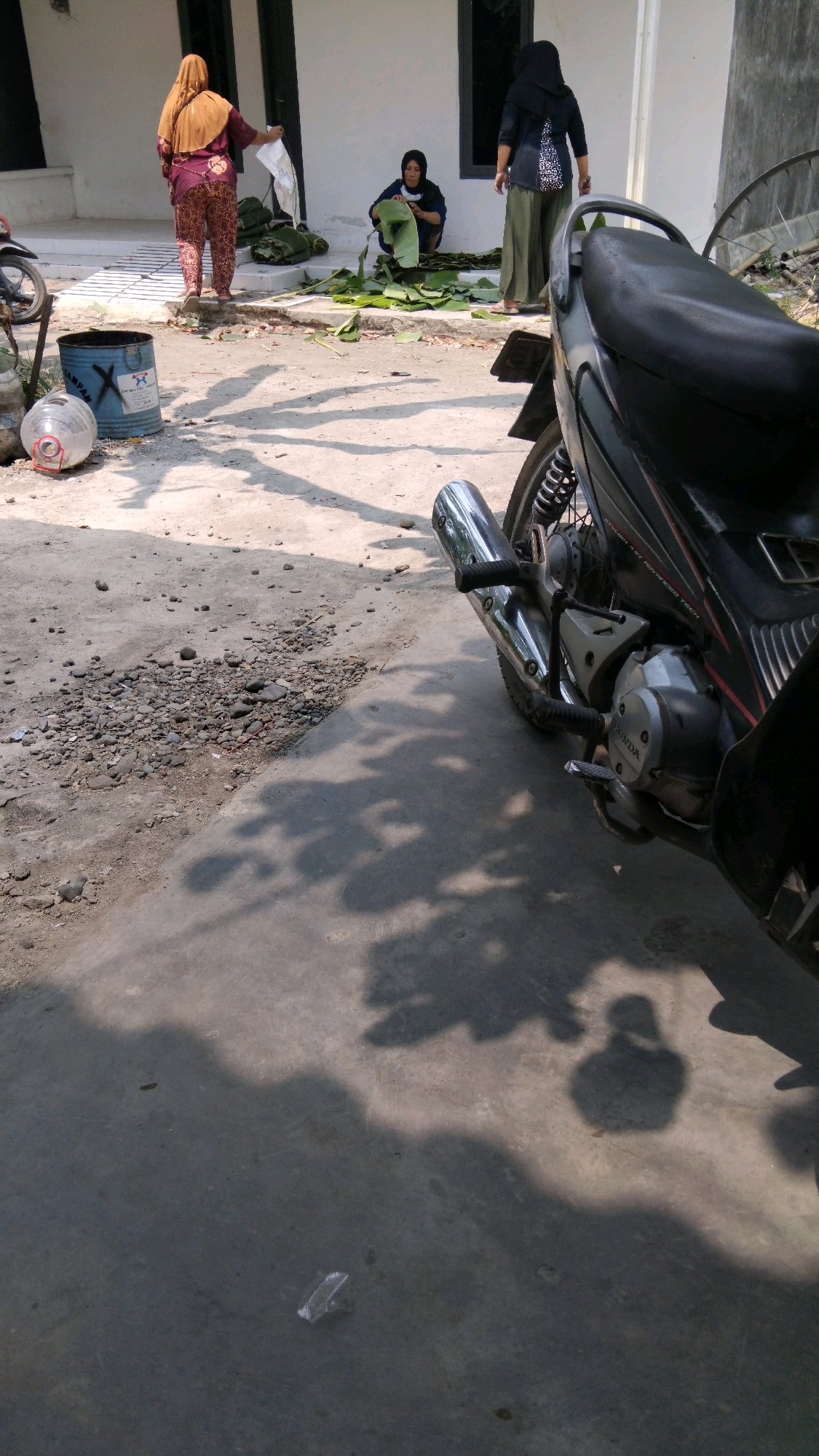 The image depicts a sunlit outdoor scene, likely in Tegal, Indonesia, with three individuals engaged in preparing food or packages using large leaves. In the foreground, the rear portion of a black motorcycle is visible. To the left, a blue barrel and a large plastic water bottle are present on the ground. The ground itself is a mix of concrete and dirt, with scattered pebbles and debris. In the background, a white building with dark-trimmed windows forms the backdrop. One woman, wearing a maroon and patterned outfit and a tan headscarf, stands near the building, holding a white sheet. Another woman, in a dark hijab and a patterned top with green pants, is seated on the ground, actively working with the leaves. A third woman, in a black hijab and a dark top with a white pattern, stands to the right, facing away from the camera. Numerous green leaves are spread out on the ground and stacked in bundles, suggesting an ongoing preparation process. Sunlight casts distinct shadows of the people and the motorcycle onto the ground, indicating a bright, sunny day.