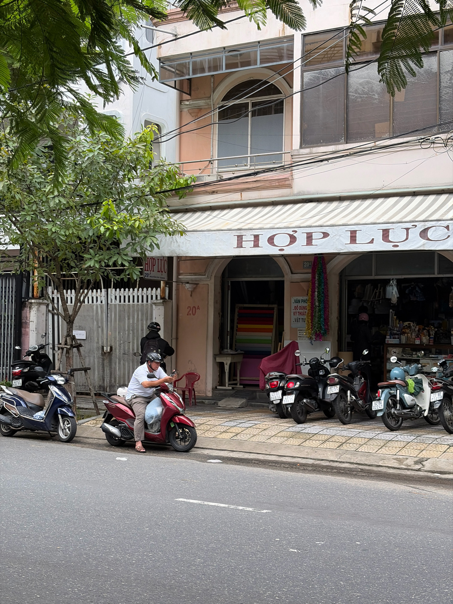 This is a medium-shot, eye-level, slightly downward-angled photograph of a street scene in Da Nang, Vietnam. In the foreground, a gray asphalt road with a white dashed line is visible. A tiled sidewalk runs alongside the road, bordered by a curb.
On the sidewalk, several parked motorcycles are lined up. The closest motorcycle is a dark red scooter, ridden by a man in a white short-sleeved shirt and gray pants. He is wearing a helmet and looking at his phone. To his left, a dark blue scooter is parked. Further down the sidewalk, more motorcycles are visible, including a black scooter and a light blue and white scooter.
Behind the motorcycles is the facade of a building. A striped awning, white with dark stripes, extends over the storefront. The building has a light pink facade and appears to be a shop. Text on the awning reads "HỢP LỤC". Below the awning, the shop entrance is visible, with merchandise displayed inside. Some hanging colorful decorations are suspended in the doorway. To the left of the shop entrance, a white fence and a tree with green foliage are present. A sign with Vietnamese text and "20A" is posted on the building. In the background, more of the building's upper floors are visible, with windows and balconies. Power lines are strung across the facade. The overall scene depicts a typical urban street in Vietnam with commercial activity and transportation.