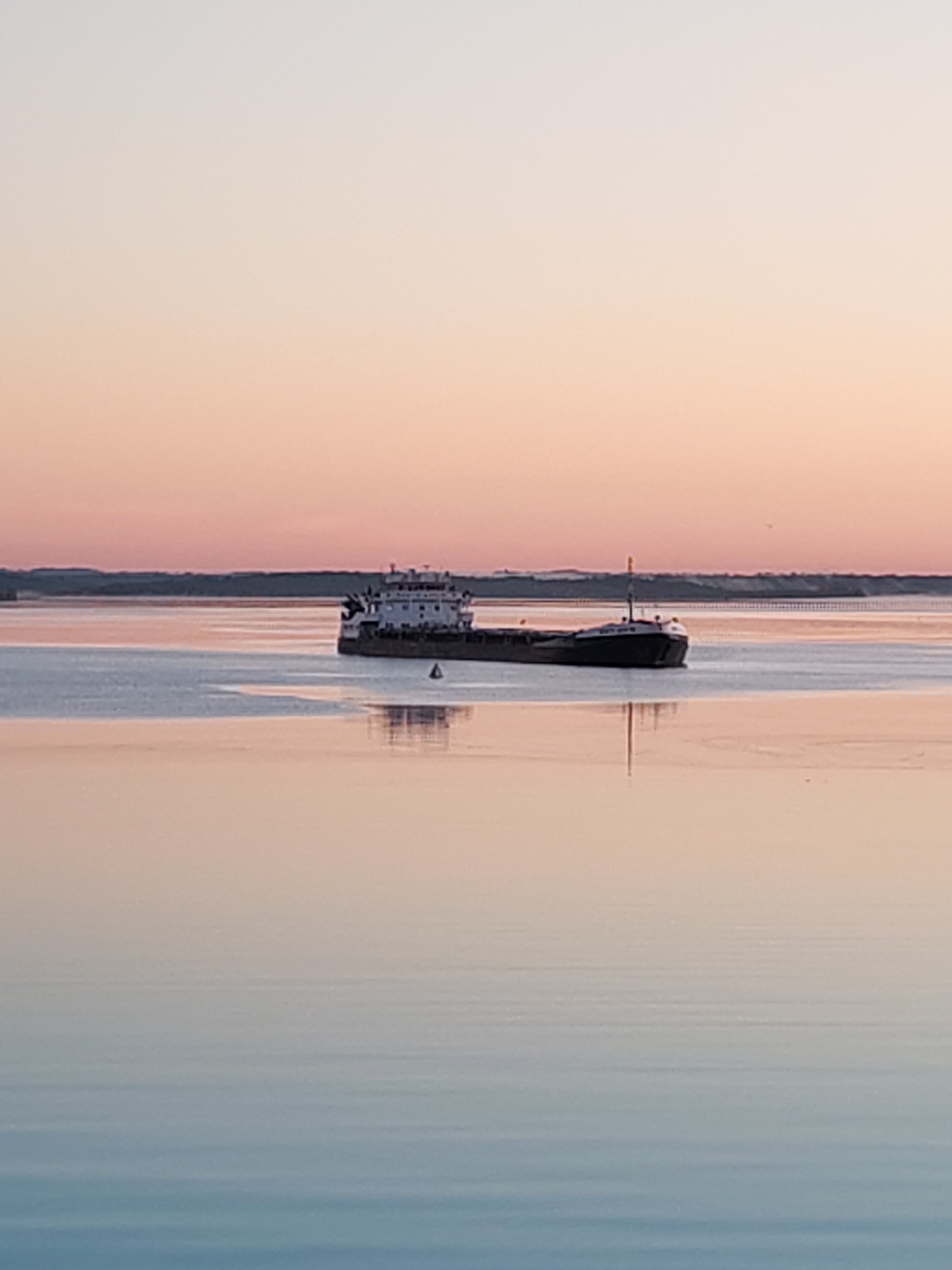 This image depicts a serene maritime scene at either sunrise or sunset, characterized by soft, diffused light.
**Location:** Unknown, Unknown
**Scene Description:**
The upper portion of the image reveals a gradient sky, transitioning from a pale peach-pink hue near the horizon to a very light, almost white-grey at the top. This gentle color gradient is indicative of dawn or dusk. A faint, dark landmass or coastline stretches across the distant horizon, with subtle, indistinguishable structures possibly visible.
The mid-ground is dominated by a large, dark-hulled cargo ship or barge, positioned slightly to the right of the vertical center. The vessel has a prominent white superstructure situated towards its stern, featuring multiple decks and discernible windows. A slender mast or pole with a small yellow or orange marker is visible at the ship's bow. A dark, triangular buoy or navigational marker floats in the water to the left and slightly forward of the ship.
The water surface is exceptionally calm and highly reflective, mirroring the sky's soft color palette. The reflection of the ship is clearly visible directly beneath it, showing minor distortion due to subtle ripples on the water. The reflective surface of the water transitions from peach-pink near the horizon to a soft, light blue-grey in the foreground. No individuals are present in the frame. The overall environment suggests a tranquil waterway.