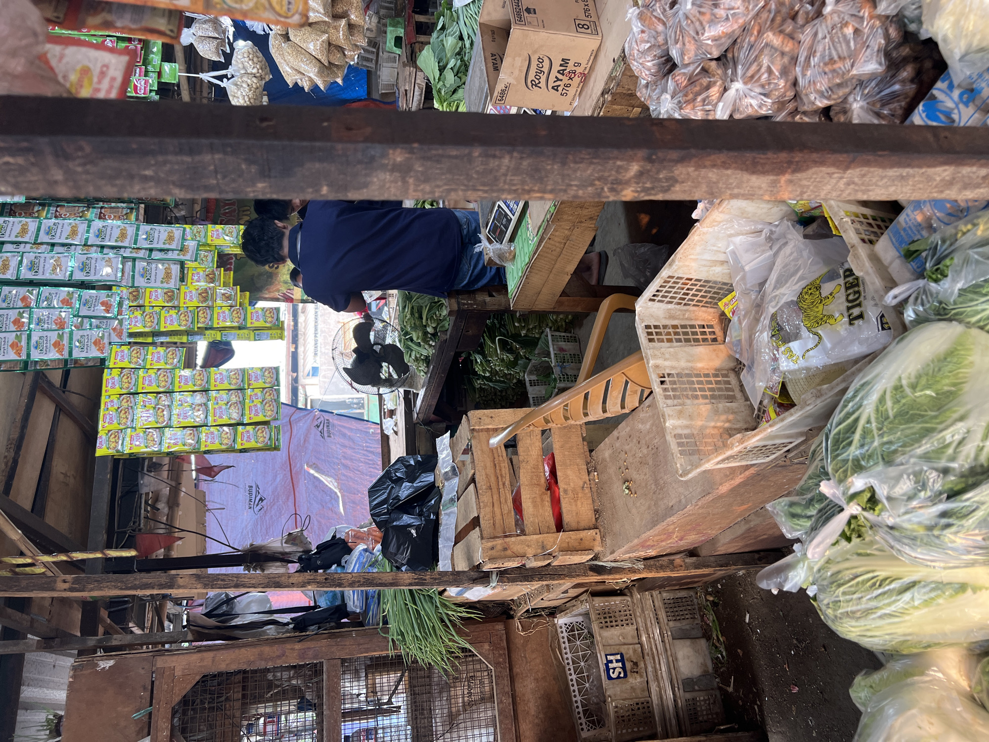 An outdoor market stall in Citeureup, Indonesia, displays a variety of produce and packaged goods. In the foreground, large cabbages are bagged in clear plastic. To the left, a white plastic crate contains a few bagged items, including one with a tiger graphic and the word "TIGER". Behind this, more plastic crates are stacked, with a larger wooden crate holding black trash bags and green onions. Further back, a man in a blue shirt and jeans stands behind a table with a scale, facing away from the camera, attending to customers. A fan is visible to his left. The stall's structure consists of rough wooden posts and beams. Shelves above are laden with rows of small, colorful packets of spices and seasonings. Bags of nuts and other dry goods hang from the ceiling. The ground appears to be dirt and debris. In the background, a blue tarp and other market stalls are visible.
