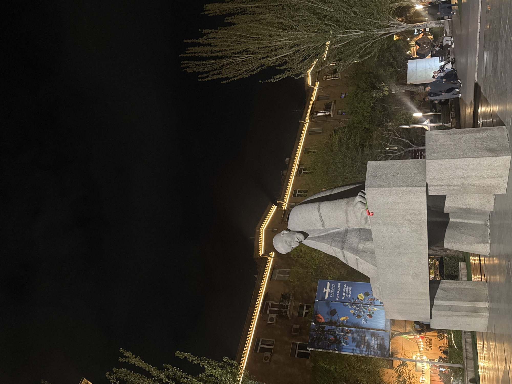 The image depicts a statue of Soviet Armenian architect Alexander Tamanian in Yerevan, Armenia, at night. The statue is made of gray stone and is positioned in front of a building adorned with lights. Banners for the Cafesjian Center for the Arts are seen near the base of the statue. People are visible in the background. The environment is urban with trees and a street.