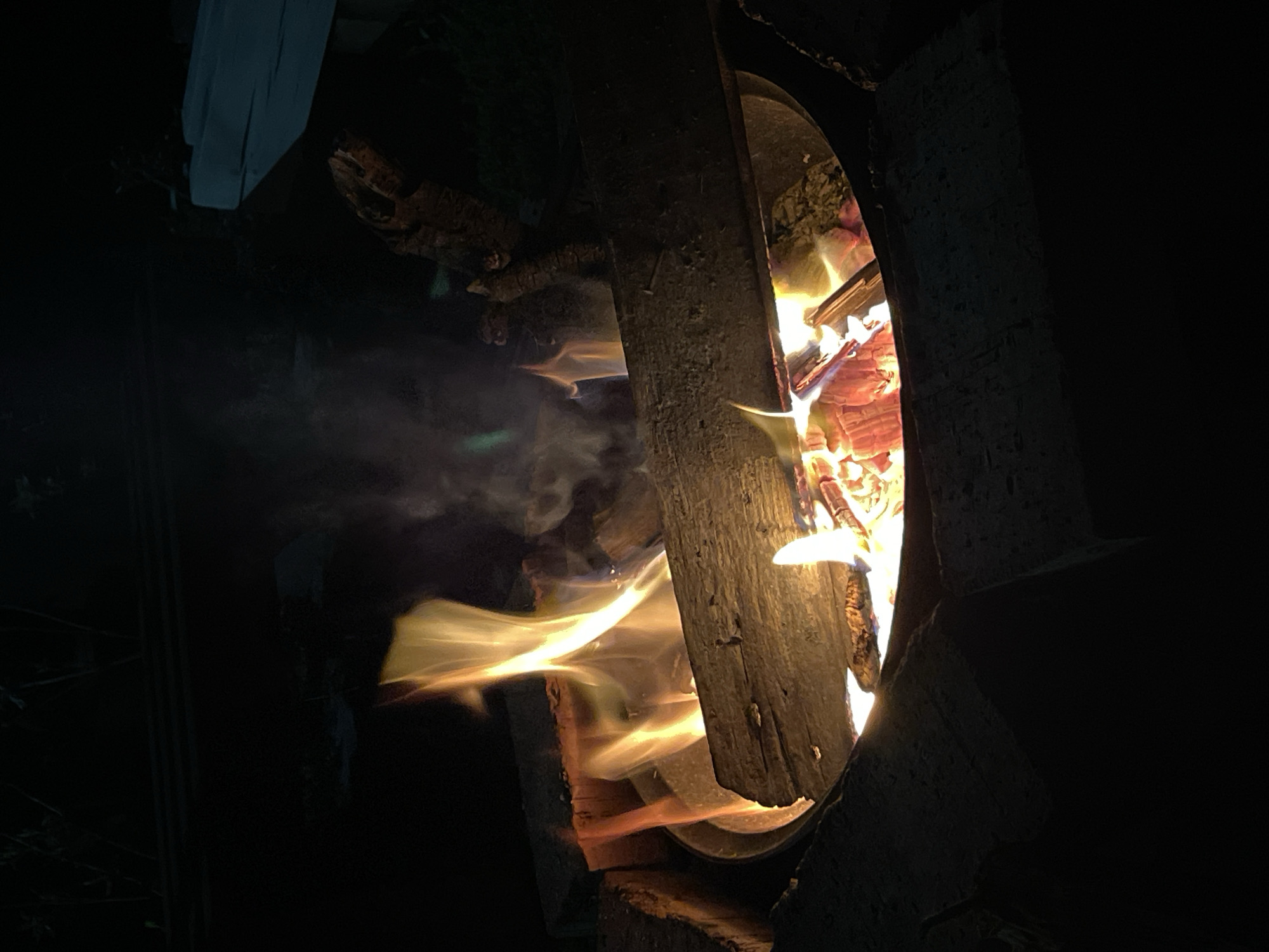 In an outdoor, dark environment, a close-up shot captures a fire pit ablaze. The fire pit is a circular metal basin, elevated by bricks on the sides. Inside the basin, logs and embers glow with fiery orange and red hues. Tall, bright yellow flames lick upward, interspersed with wisps of smoke. A large, rough-hewn piece of wood lies horizontally across the fire pit, partially obscuring the flames. The background is dark, with faint outlines of foliage and a wooden structure visible. The scene suggests a nighttime gathering around a campfire.