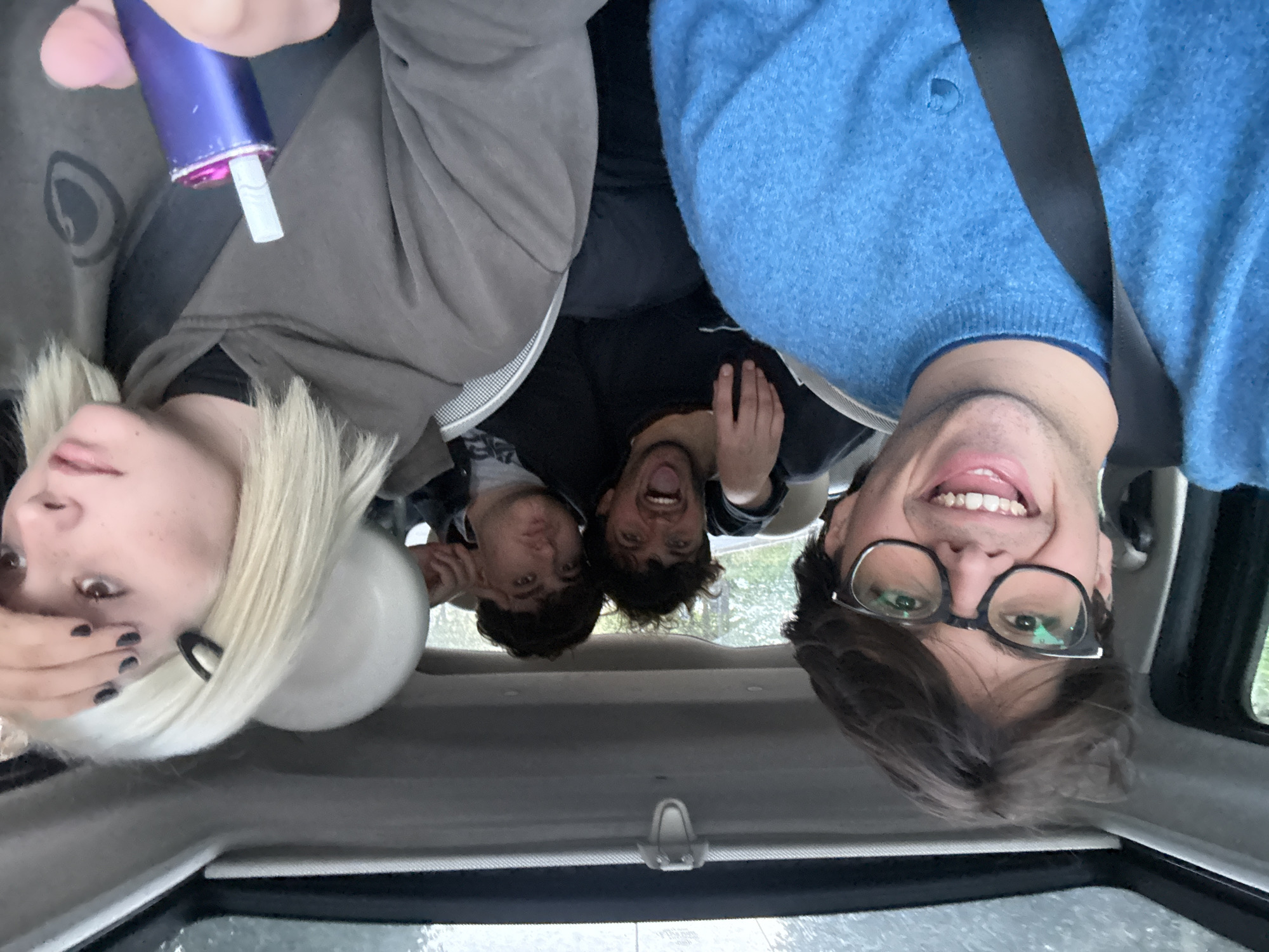 Five individuals are visible inside a car. The perspective suggests the photo was taken from the roof of the vehicle.
From left to right:
Person 1: A light-skinned individual with bleached blonde hair and dark nail polish. They appear to be holding a purple electronic cigarette device.
Person 2 and 3: Two darker-skinned individuals in the back. They are huddled together.
Person 4: A light-skinned individual wearing a blue sweater and a seatbelt.
A white balloon floats near the first person.
The car's interior is beige. Rain is visible on the car window. The location is not discernible from the image. The city of Milan cannot be identified.