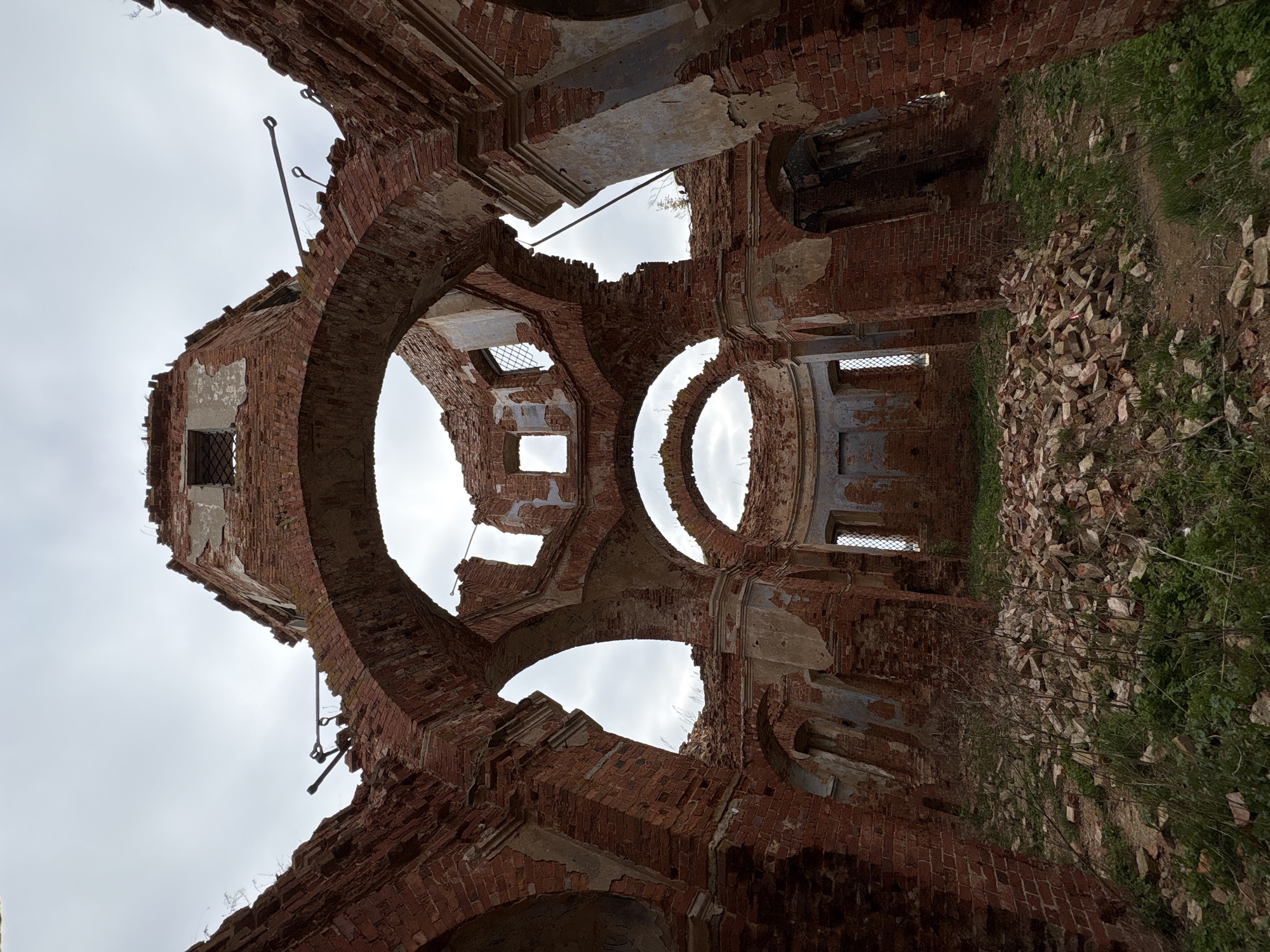 The image captures an upward-looking view from within the heavily ruined interior of a large brick structure, likely an old church or cathedral, in Staroe Rakomo, Russia. The roof and significant portions of the upper walls have collapsed, revealing an overcast, grey sky. The scene is dominated by a complex network of crumbling, weathered red brick arches and circular openings that once supported a dome or ceiling. Patches of faded plaster cling to some brick surfaces, indicating former decorative finishes. Several rusty metal rods, possibly remnants of structural reinforcements, protrude from the damaged brickwork. At the base of the visible walls, piles of fragmented bricks and debris are scattered across the ground, partially overgrown by green grass and sparse vegetation. Some smaller rectangular window openings with remnants of grates are visible in the lower sections of the walls. No people are present, no specific activity is taking place, and there is no discernible text. The diffused light from the cloudy sky contributes to a somber and desolate atmosphere, highlighting the advanced state of decay.