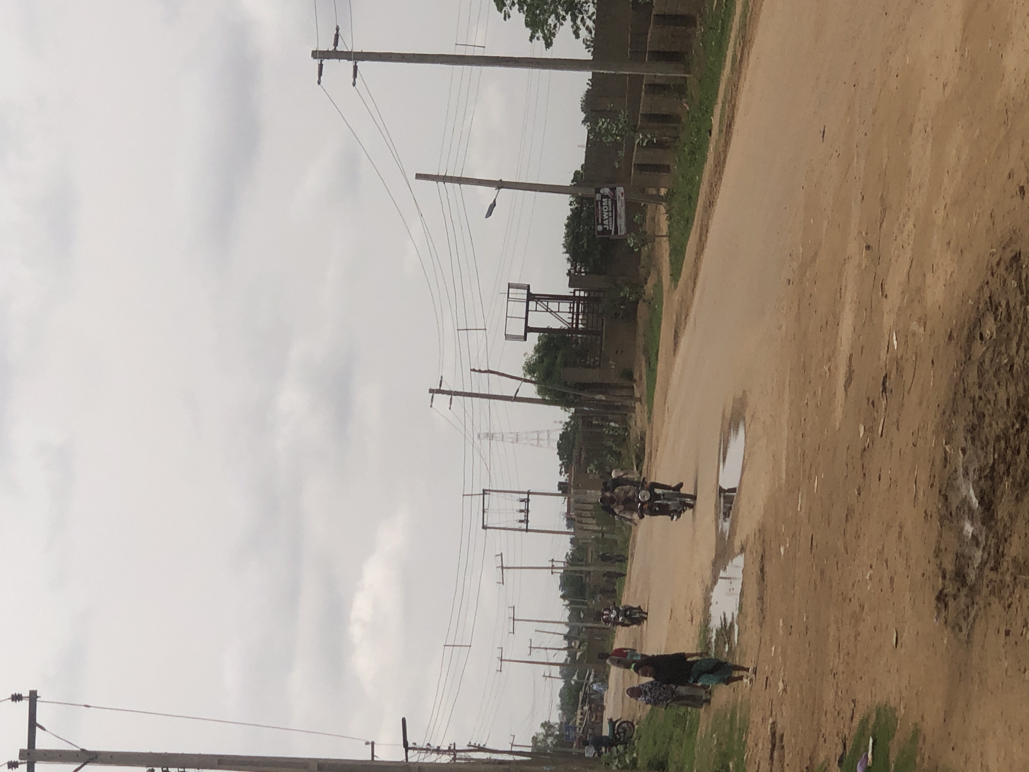 The image shows a dirt road in Hanwa, Nigeria. Several people and motorcycles are on the road. Overhead power lines run along the roadside. Some puddles of water are visible on the road. The sky is overcast. Buildings and vegetation are visible on the side of the road. A metal structure resembling a water tower can be seen further down the road.