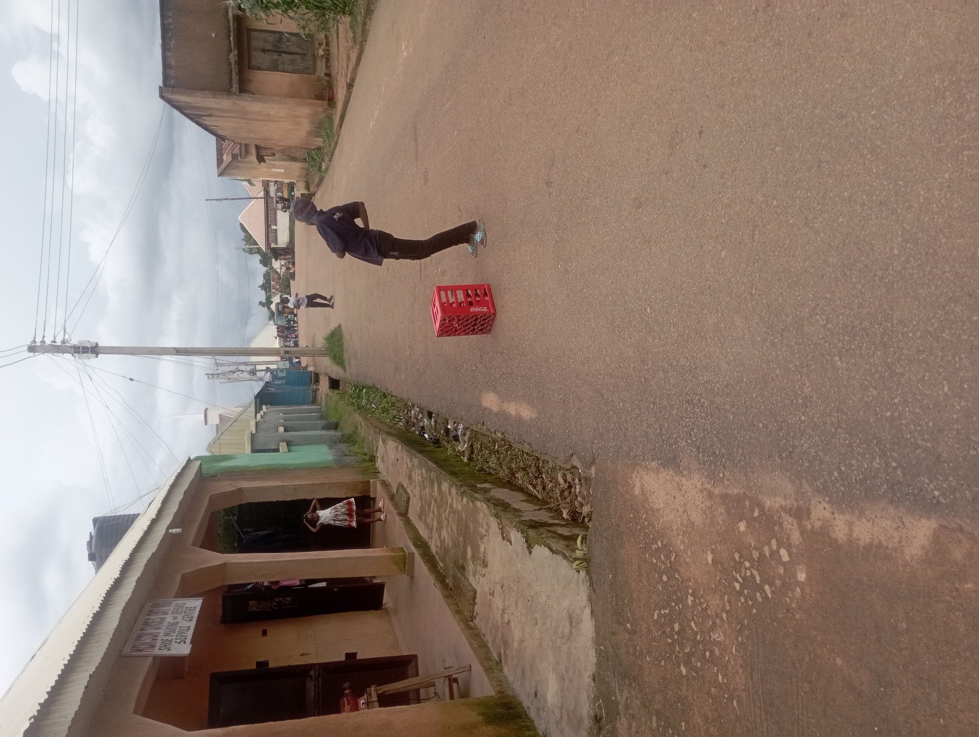 The image captures a street scene in Rafin Bauna, Nigeria, under an overcast sky. On the left, a storefront displays a sign that reads "MOISON SHOES DIY. MBO SHOE MAKING AND REPAIRS SERVICE CENTRE". A young girl in a patterned dress stands in the doorway of the shop. In the middle ground of the street, a red Coca-Cola crate rests on the asphalt. A young person wearing a dark hoodie and black pants is bent over with their back arched, looking towards the crate. Further down the street, two other figures are visible, one walking and another standing. The environment includes a paved road, a concrete drainage ditch with debris, and buildings on either side of the street.