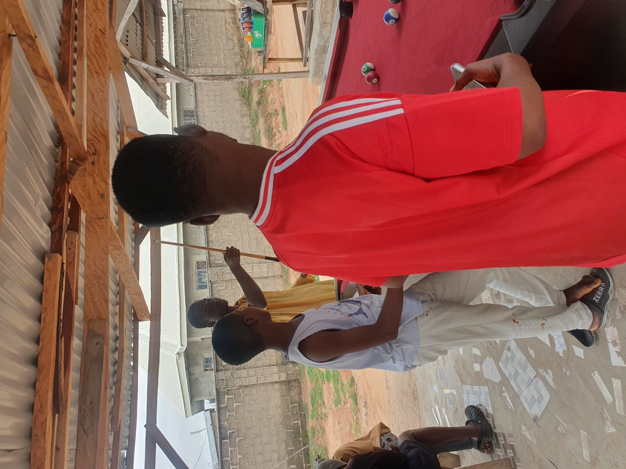 In Lokoja, Nigeria, under a corrugated metal roof supported by rough-hewn wooden beams, three young men are gathered around a red pool table. One man, wearing a yellow garment, stands with a pool cue, appearing to instruct or engage with the others. A second young man, in a grey tank top and white trousers, stands to his left, looking towards the pool table. The third, viewed from behind and wearing a bright red t-shirt with white stripes on the sleeves, holds what appears to be a smartphone in his right hand. Pool balls are scattered on the table, indicating a game is in progress or has recently concluded. The environment suggests an outdoor or semi-outdoor recreational space, with a brick wall in the background and unpaved ground.