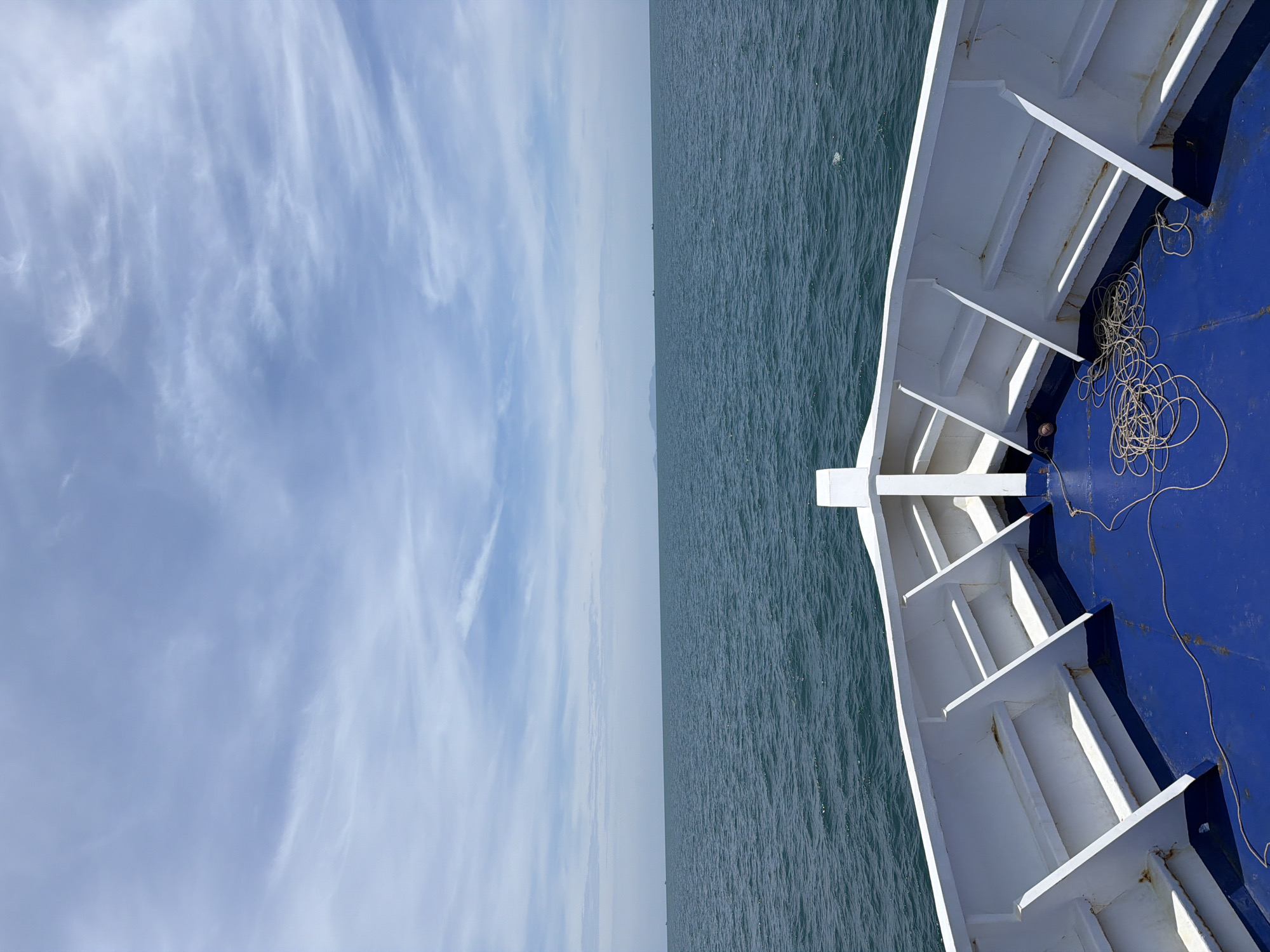 The image shows a view from the side of a boat on the water, under a cloudy sky. The boat has a blue deck with white structural supports. There is coiled rope on the deck. The water stretches to the horizon. The sky is light blue with wispy white clouds. The location is likely near ตำบลเกาะพะงัน, Thailand, given the prompt.