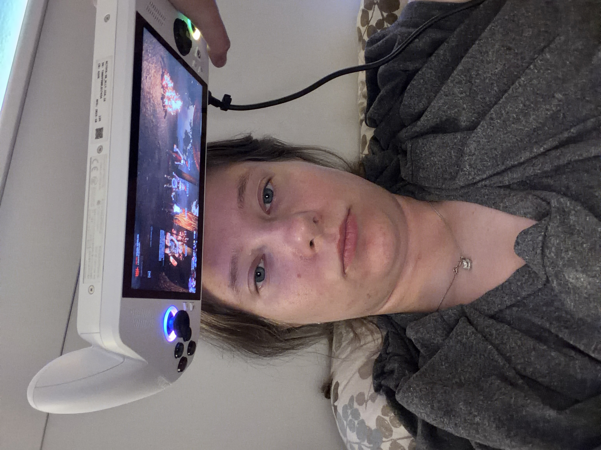 The image shows a person holding a white gaming device above their head. The device has a screen displaying a video game, game controls, and a cable attached to it. The person is lying down, with their head resting on a pillow. The person is wearing a gray shirt and a necklace. The background is a wall. The scene appears to be in a bedroom. There is no information about the city of Portland, United States of America, in this image.