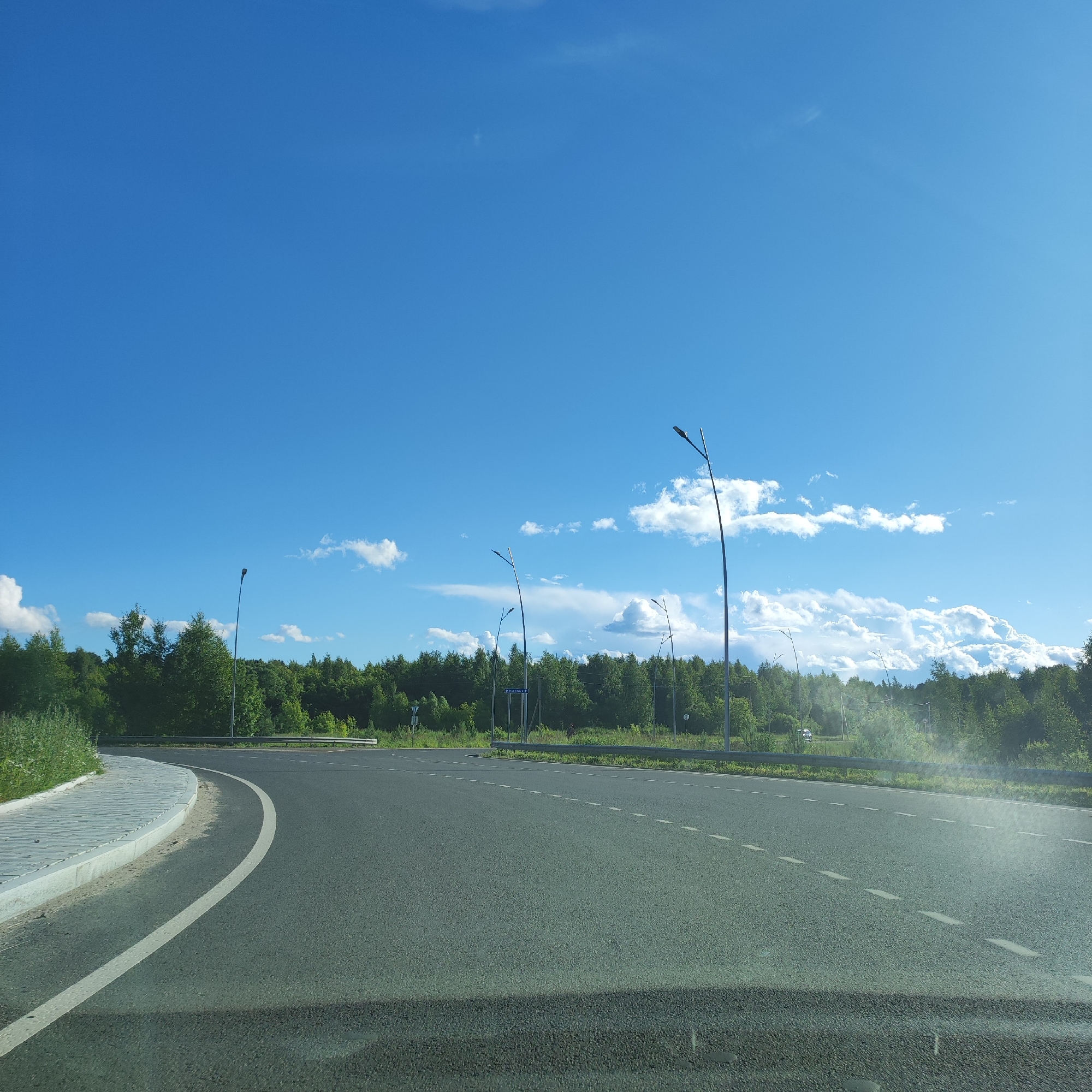 This is a wide-angle, eye-level shot of a paved road curving to the left. The road is marked with dashed white lines. On the left side of the road, there is a paved sidewalk with a concrete curb. Beyond the road and sidewalk, a dense line of green trees forms the background, with a few streetlights interspersed among them. The sky above is a vibrant, clear blue with scattered white, fluffy clouds, suggesting a sunny day.
The scene appears to be a road on the outskirts of a town or in a rural area, possibly a highway exit or entrance, given the presence of streetlights and the wide expanse of road. The lush greenery indicates a warm season, and the bright sky points to daytime, likely late morning or afternoon due to the sunlight. There are no people or animals visible in the frame.
A blue road sign with white text is visible in the middle ground, just beyond the streetlights. The text on the sign is difficult to read clearly but appears to be directional. There are also some faint markings on other signs and possibly some distant structures or vehicles obscured by the trees and distance. The overall impression is one of an open, tranquil landscape on a pleasant day. The location context is Октябрьский, Russia.
