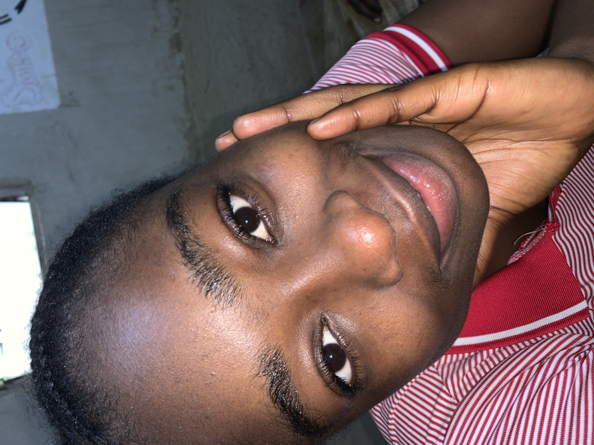 This is a close-up, portrait-style photograph of a young Black woman. She is looking directly at the camera, with her left hand resting on the right side of her face, obscuring part of her cheek and ear. Her eyes are dark brown and open, and she has a slight smile. Her lips are closed and appear to be glossy. She has dark, short hair styled in braids. She is wearing a red and white striped collared shirt. The background is out of focus and appears to be an indoor environment with a concrete wall that has some graffiti on it and a window in the upper left corner. The lighting suggests an indoor setting, possibly at dusk or dawn. There is no indication of the specific location in Rafin Par, Nigeria, other than it being an indoor scene.