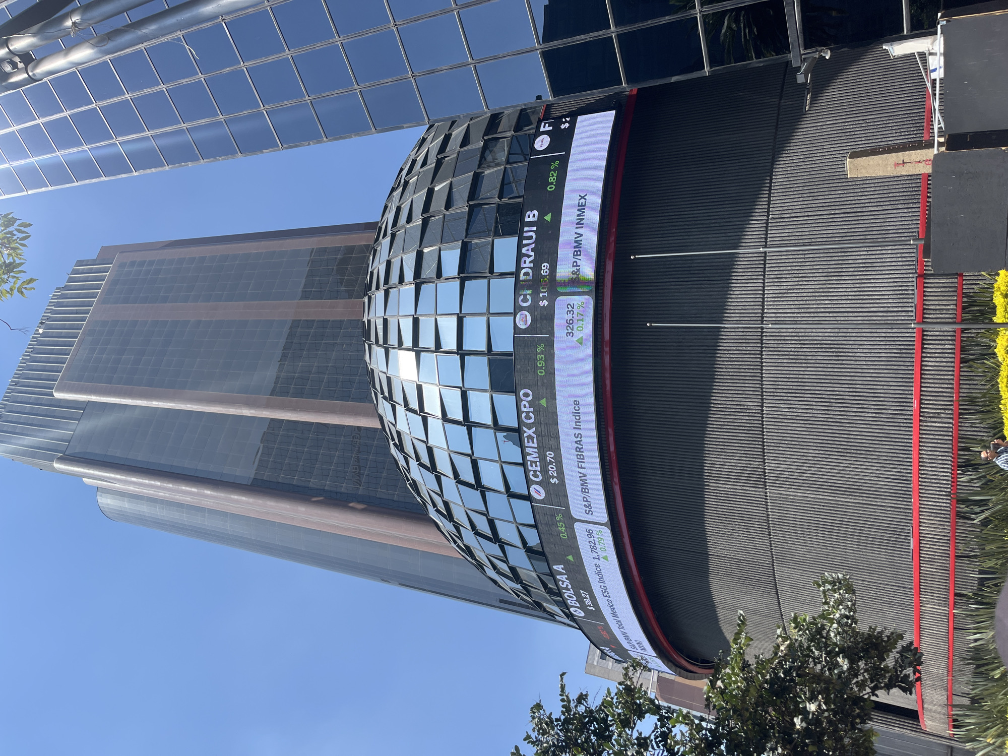 The image shows the Mexican Stock Exchange (Bolsa Mexicana de Valores) in Mexico City, Mexico. The iconic rounded structure covered in mirrored squares is visible, as well as a large LED ticker displaying financial information for various companies and indices. The ticker shows values for BOLSA A, CEMEX CPO, CHIDRAUI B, S&P/BMV indices, and others. A modern skyscraper rises in the background. In the foreground, there is some greenery. The sky is clear and blue.