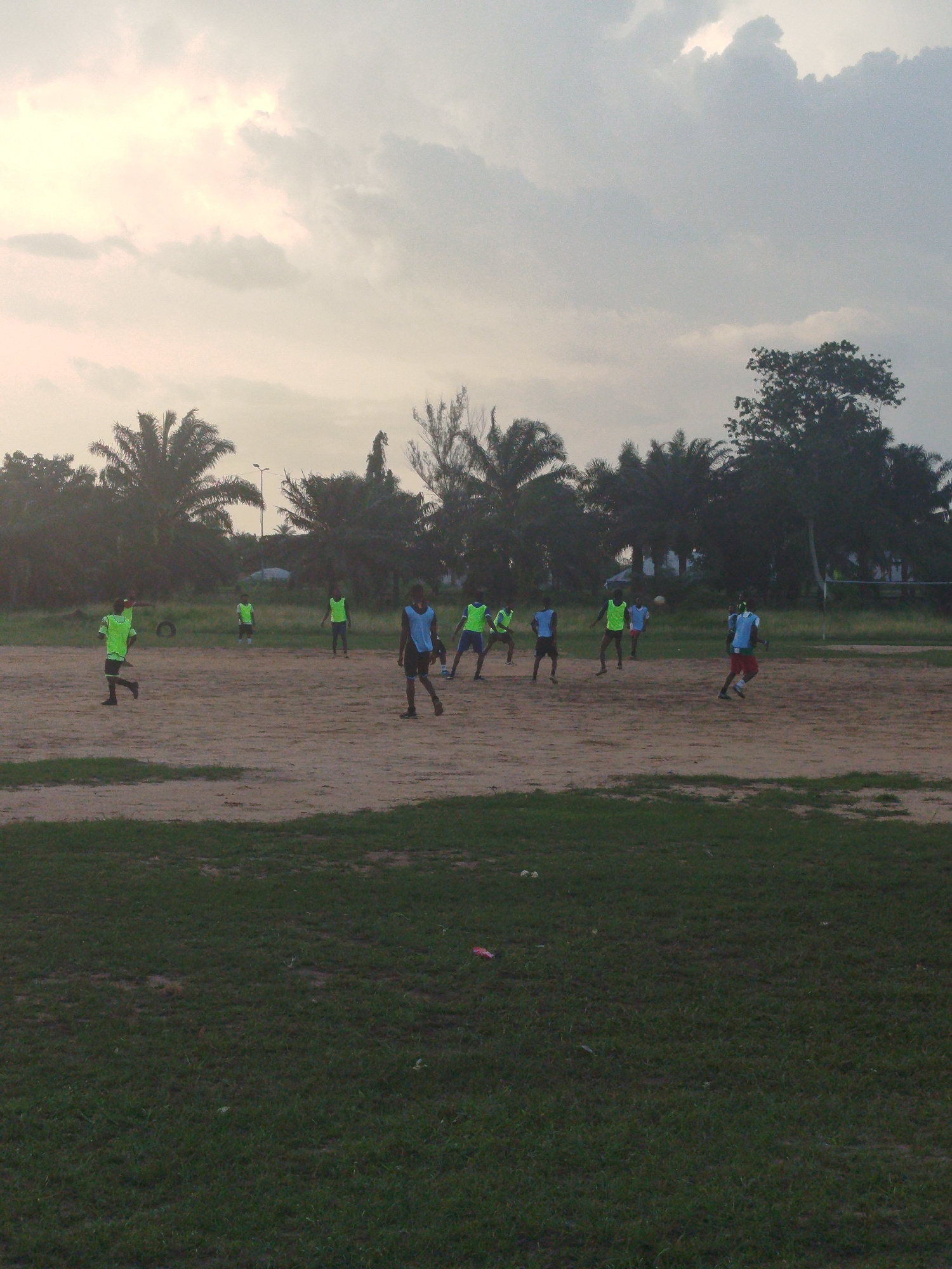 The scene depicts a group of young men playing soccer on a dirt field in Ikot Mbonde, Nigeria. The field is bordered by palm trees and other vegetation, suggesting a location on the outskirts of the city. The sky is overcast, indicating late afternoon or early morning lighting. The soccer players are wearing a mix of colored shirts, possibly used as team identifiers. Several players are wearing green vests over their shirts, while others wear blue vests. The foreground features patches of grass leading to the playing field, suggesting a transition from a more maintained area to a more natural, unpaved space. A white building is visible in the far background, and some poles are seen.