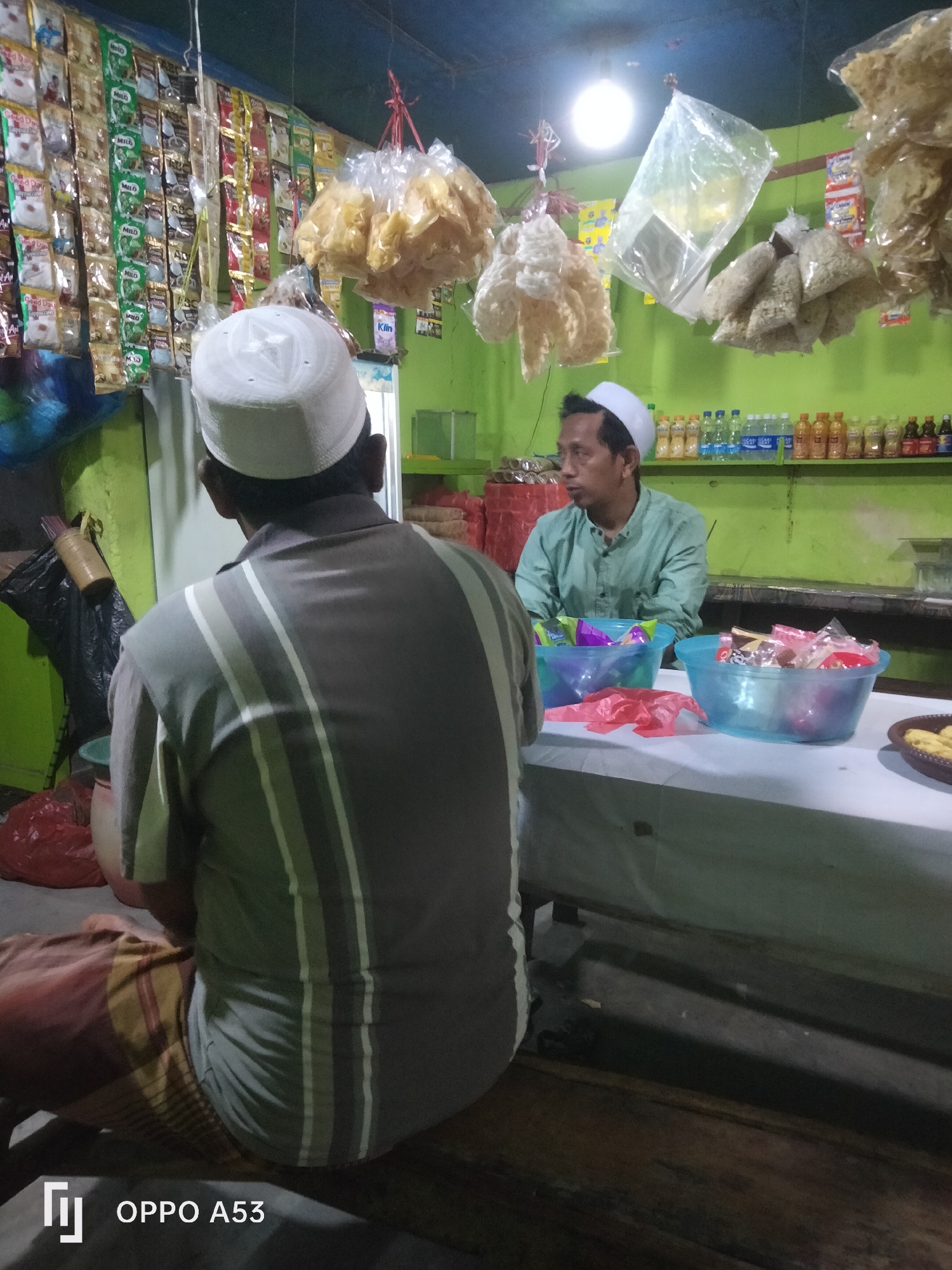 This image depicts the interior of a small shop, possibly a warung or minimarket, in Badung, Indonesia. The shop appears to be dimly lit, with the primary light source a single overhead bulb. The walls are painted a vibrant green. Shelves line the back wall, stocked with bottled beverages and various packaged goods. Hanging from the ceiling are bags of snacks, including what appear to be puffed chips or crackers.

In the foreground, two men are visible. One man, seen from behind, is wearing a grey and white striped shirt and a white skullcap, and is seated on the floor or a low stool. His lower body is obscured by a sarong or similar cloth. The second man, positioned behind a counter, is wearing a light green long-sleeved shirt and a white skullcap. He is looking to his left, and his hands are resting on the counter in front of him. On the counter are two blue plastic bowls filled with packaged snacks, and a red plastic bag.

The overall impression is that of a local convenience store or a small food stall operating in a residential or commercial area of Badung. The presence of packaged snacks and bottled drinks suggests it serves as a place for locals to purchase everyday items.