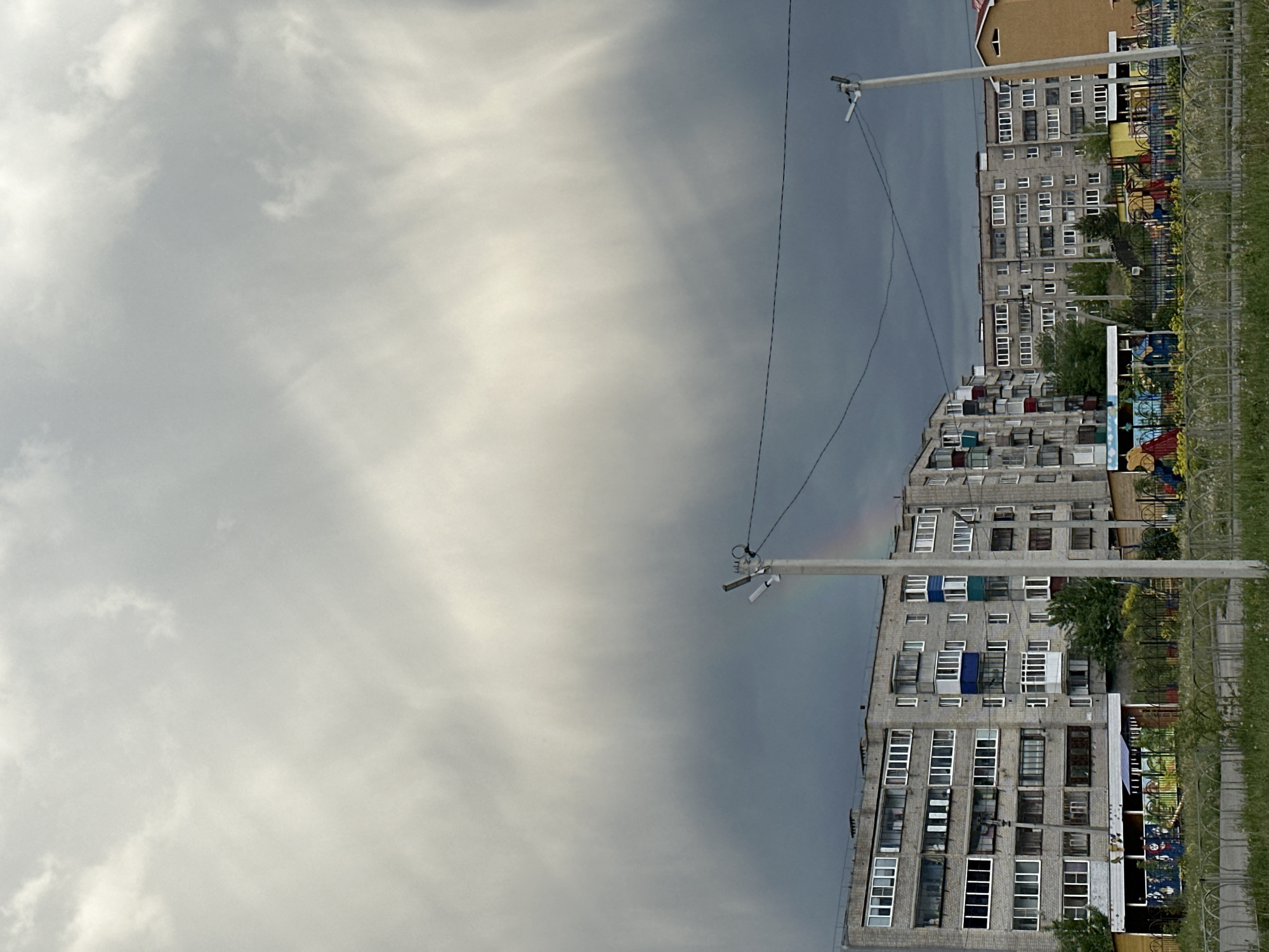 The image shows a residential area in an unidentified city. Several multi-story apartment buildings with numerous windows are visible. In front of the buildings, there is a fenced playground. Overhead power lines run across the scene, suspended by utility poles. The sky is overcast, with heavy, gray clouds dominating the view. A faint rainbow is visible near one of the power poles.
