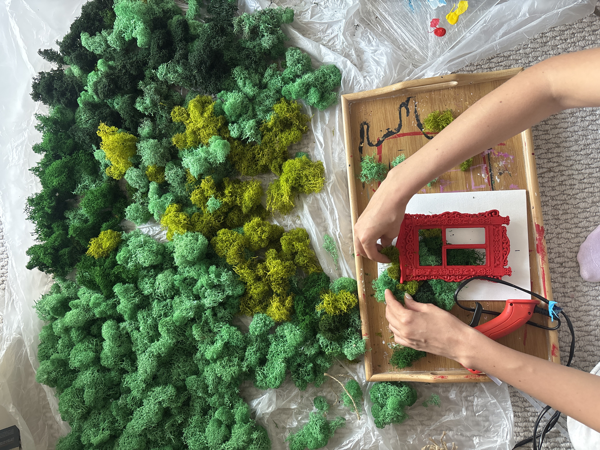 A high-angle shot shows a person arranging green and yellow moss on a wooden tray. The moss is displayed on a sheet of plastic wrap to the left of the tray. A red decorative frame and a red hot glue gun also sit on the tray. The person is using their hands to press pieces of moss onto the frame. Dabs of red, yellow, and blue paint are visible on the plastic wrap. The scene is situated on a gray carpet.
The city of Krasnoyarsk, Russia is not discernible in this image.