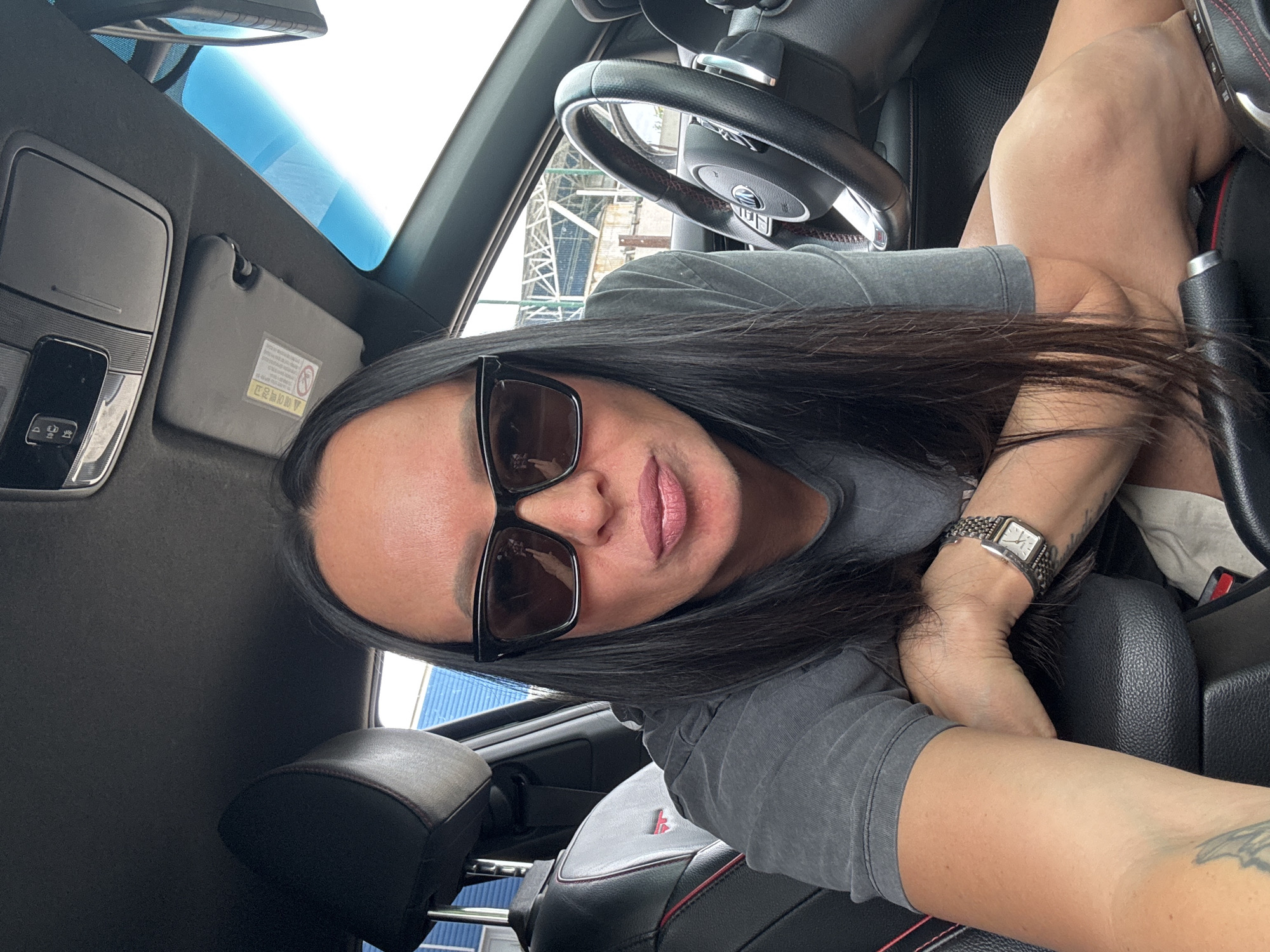 A woman is taking a selfie inside a car. She is wearing sunglasses and a gray t-shirt, and has long black hair. A watch is visible on her left wrist. The car's interior is black with red stitching on the seats and steering wheel. Through the car's windows, indistinct buildings can be seen in the background. No specific location in Chaikovsky, Russia can be determined from the image.