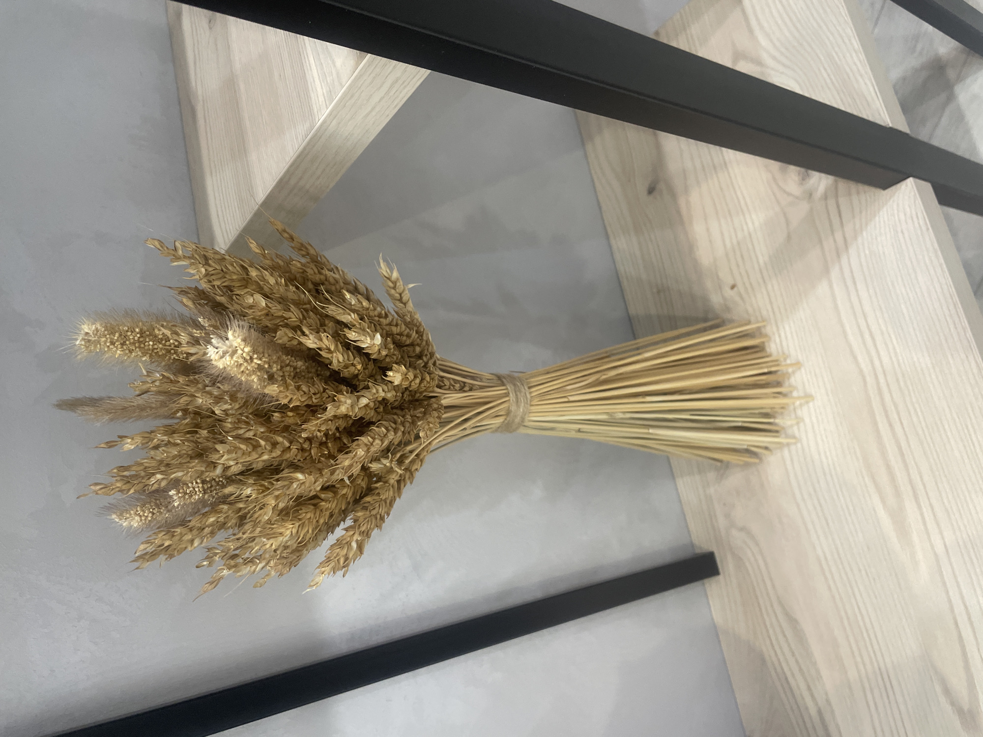 A bouquet of dried wheat stalks is displayed on a light-colored wooden shelf, which is part of a shelving unit with black metal supports. The background consists of a gray wall. The overall scene is indoors, suggesting a domestic or retail environment. No specific landmarks or indicators of Saint Petersburg, Russia, are visible.