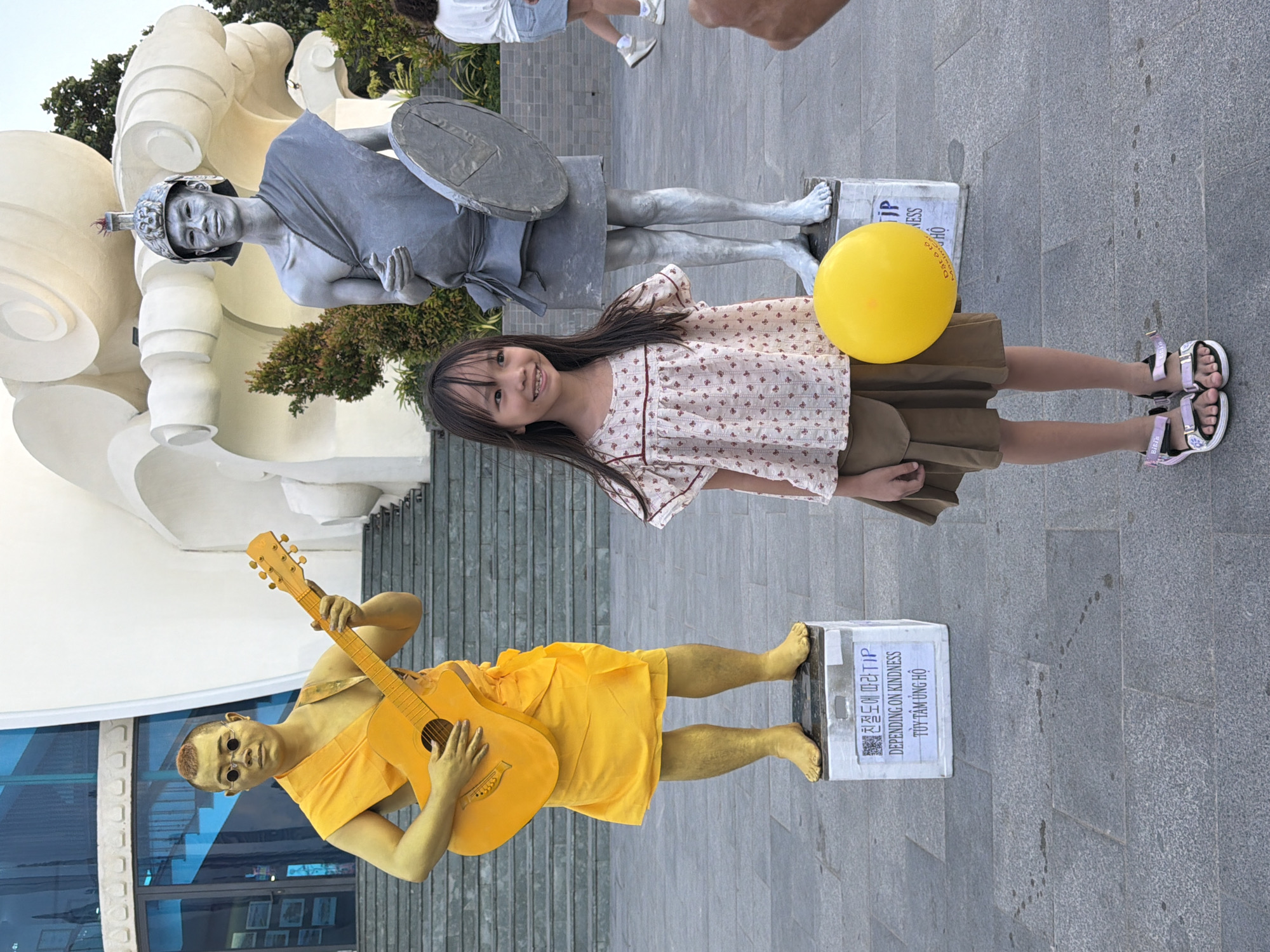 In Nha Trang Ward, Vietnam, a young girl stands smiling in front of two street performers painted gold and silver. The girl, with dark hair and wearing a floral blouse and brown skirt, holds a yellow balloon. To her left, a performer painted gold, wearing an orange robe and round glasses, holds a guitar. To her right, a performer painted silver, dressed in a grey robe and helmet, holds a shield. They are posed on pedestals in front of a modern building with glass windows and stairs. The ground is paved with grey tiles.