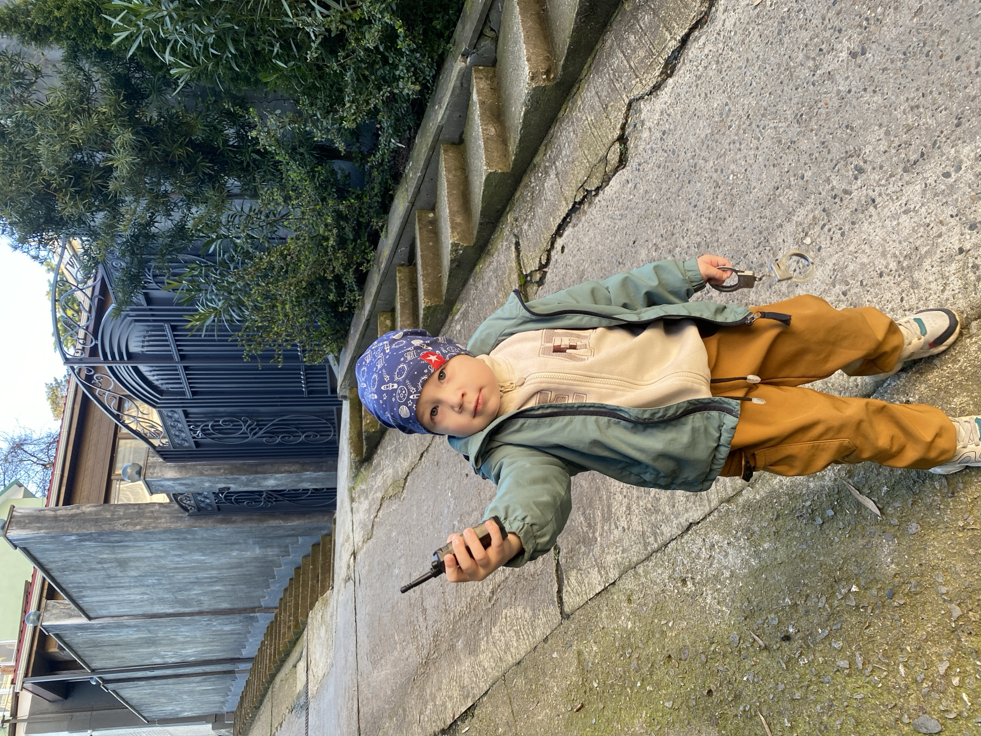 A young child stands on a concrete walkway in Sochi, Russia. The child is wearing a blue bandana, a beige sweatshirt, a green jacket, and mustard-colored pants with white and gray sneakers. The child holds a black walkie-talkie in one hand and a pair of handcuffs in the other. To the left of the child are concrete steps and a wrought-iron gate leading to a building. Lush green foliage is visible behind the gate. The concrete walkway shows signs of wear and cracking. The image is taken at a slightly tilted angle.
