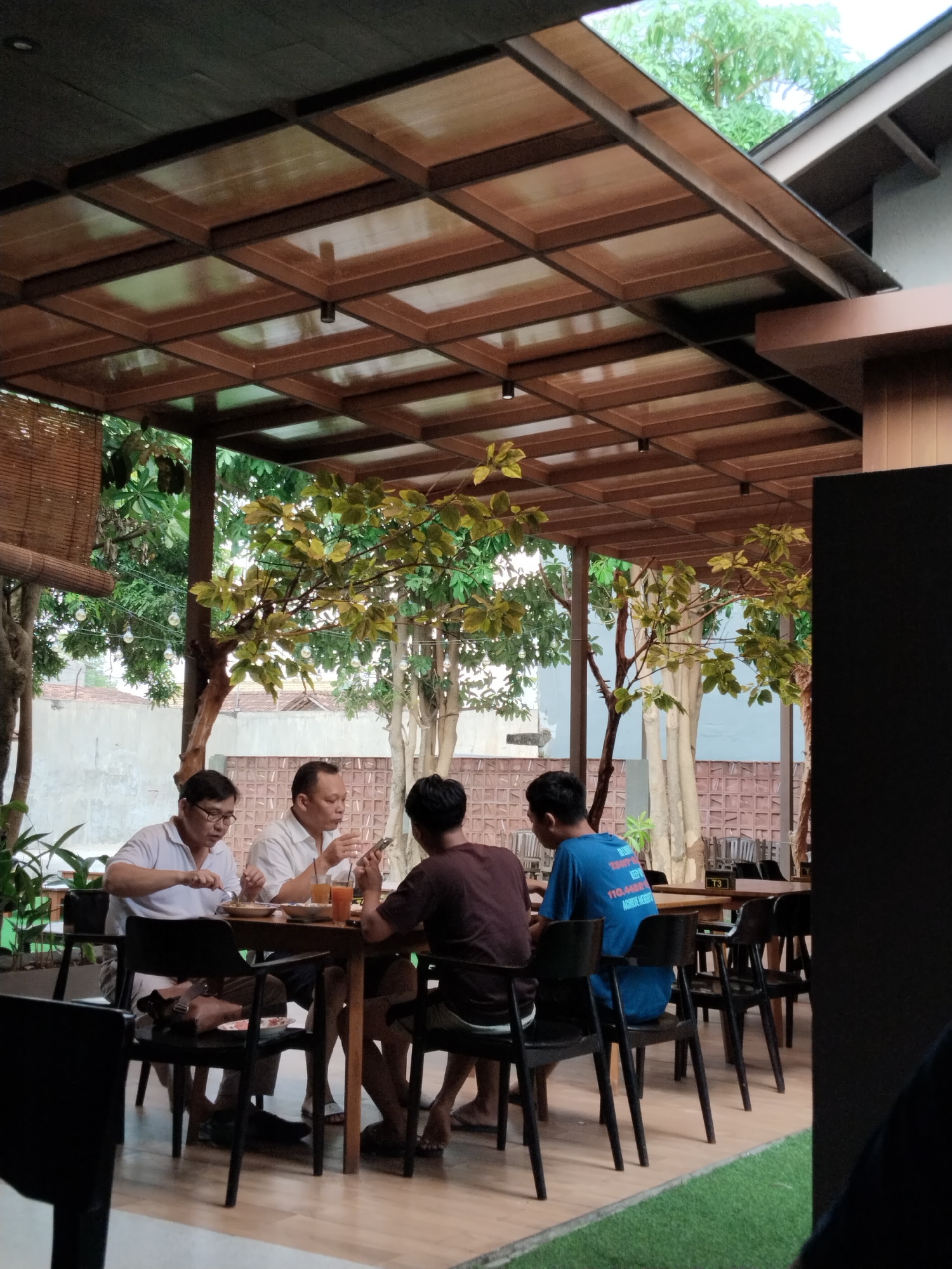 Four individuals are seated at a wooden table outside an establishment. Two men are in the foreground, with one wearing a white polo shirt and glasses, actively eating with a fork and knife. Beside him, another man, also in a white shirt, is engaged with his phone and eating. Behind them, two other individuals are seated, one in a maroon shirt and the other in a blue t-shirt with text on the back, facing away from the camera. The table is laden with plates of food, drinks in clear glasses, and cutlery. The scene is set under a wooden slatted roof structure with translucent panels, allowing natural light to filter through. Lush green foliage and trees are visible in the background, suggesting an outdoor or semi-outdoor dining area. The flooring appears to be wooden planks. The overall environment suggests a casual dining setting in Kecamatan Pati, Indonesia.