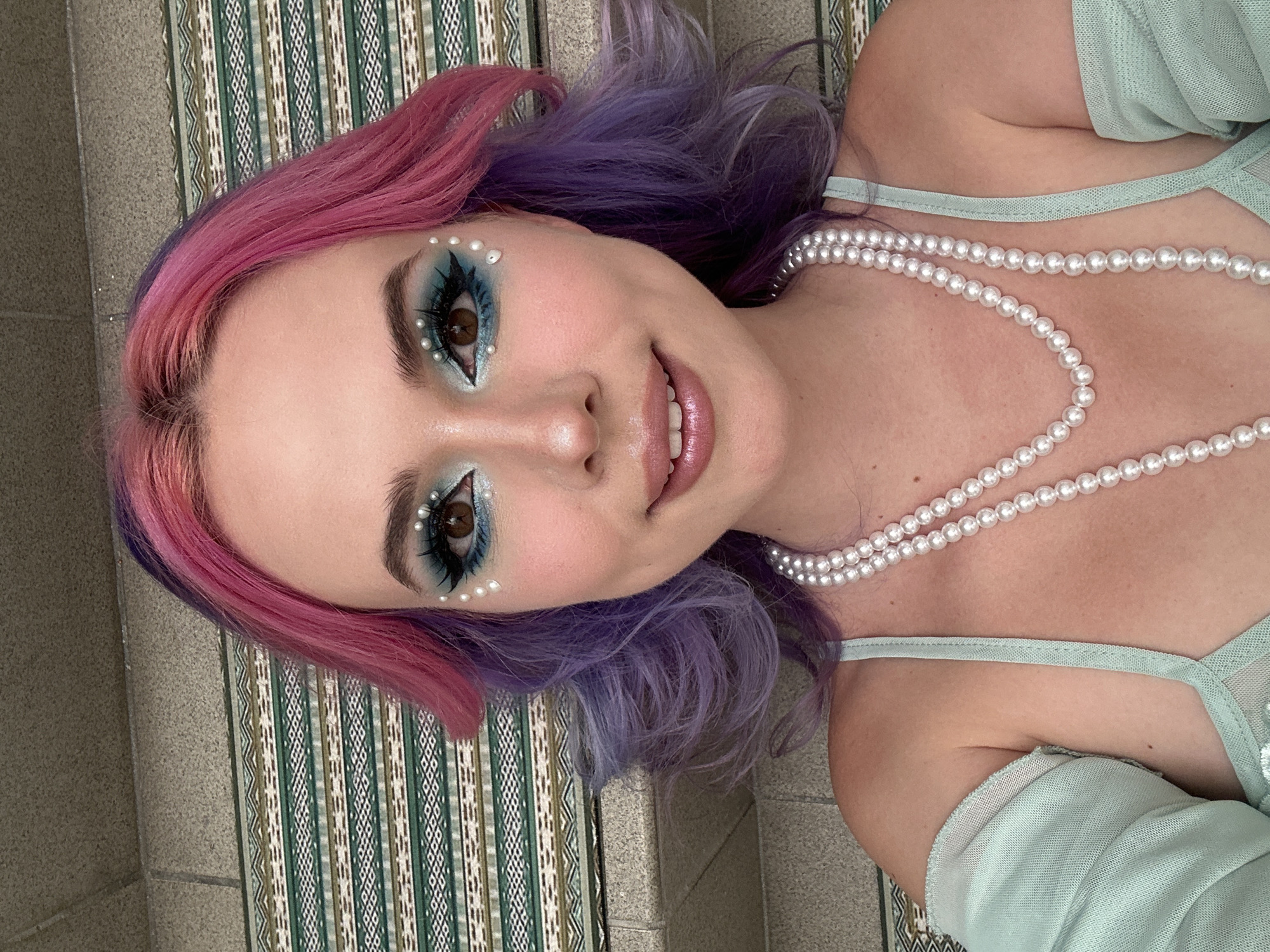 A young woman with pink and purple hair lies on a surface with a patterned rug, possibly steps, wearing light blue clothing and a double strand of pearls. She has blue eye makeup with small pearl accents and is smiling. The scene's location in the City of Zagreb, Croatia, is not discernible from the image alone.