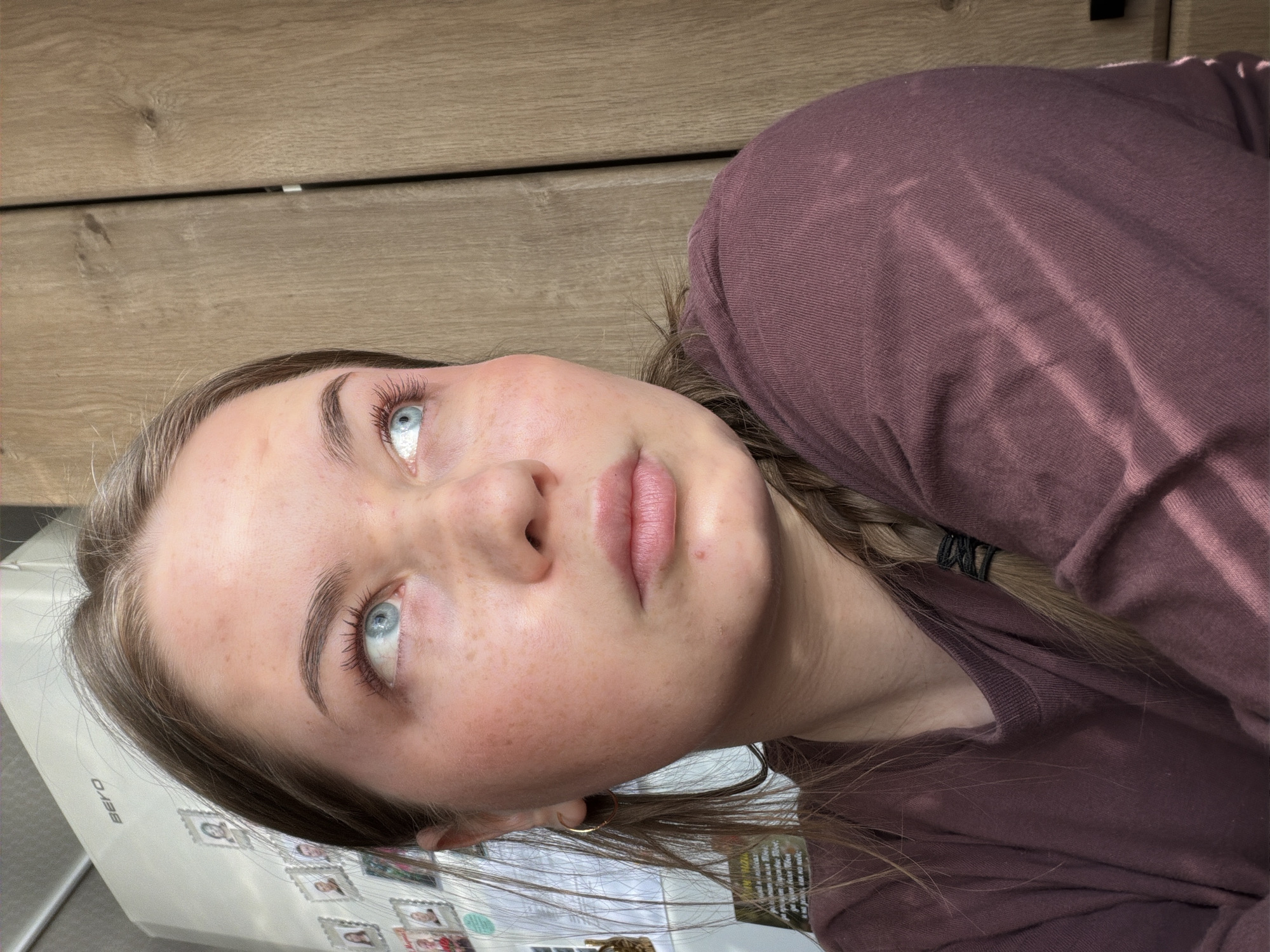A young woman with fair skin, light brown hair, and striking blue eyes is looking upwards. She has freckles and some minor skin imperfections. She is wearing a burgundy-colored long-sleeved shirt. Part of her hair is braided. The background includes what appears to be a light-colored wood cabinet and the side of a white refrigerator covered with magnets and pictures. The location of Уральское, Russia is not discernible from the image.