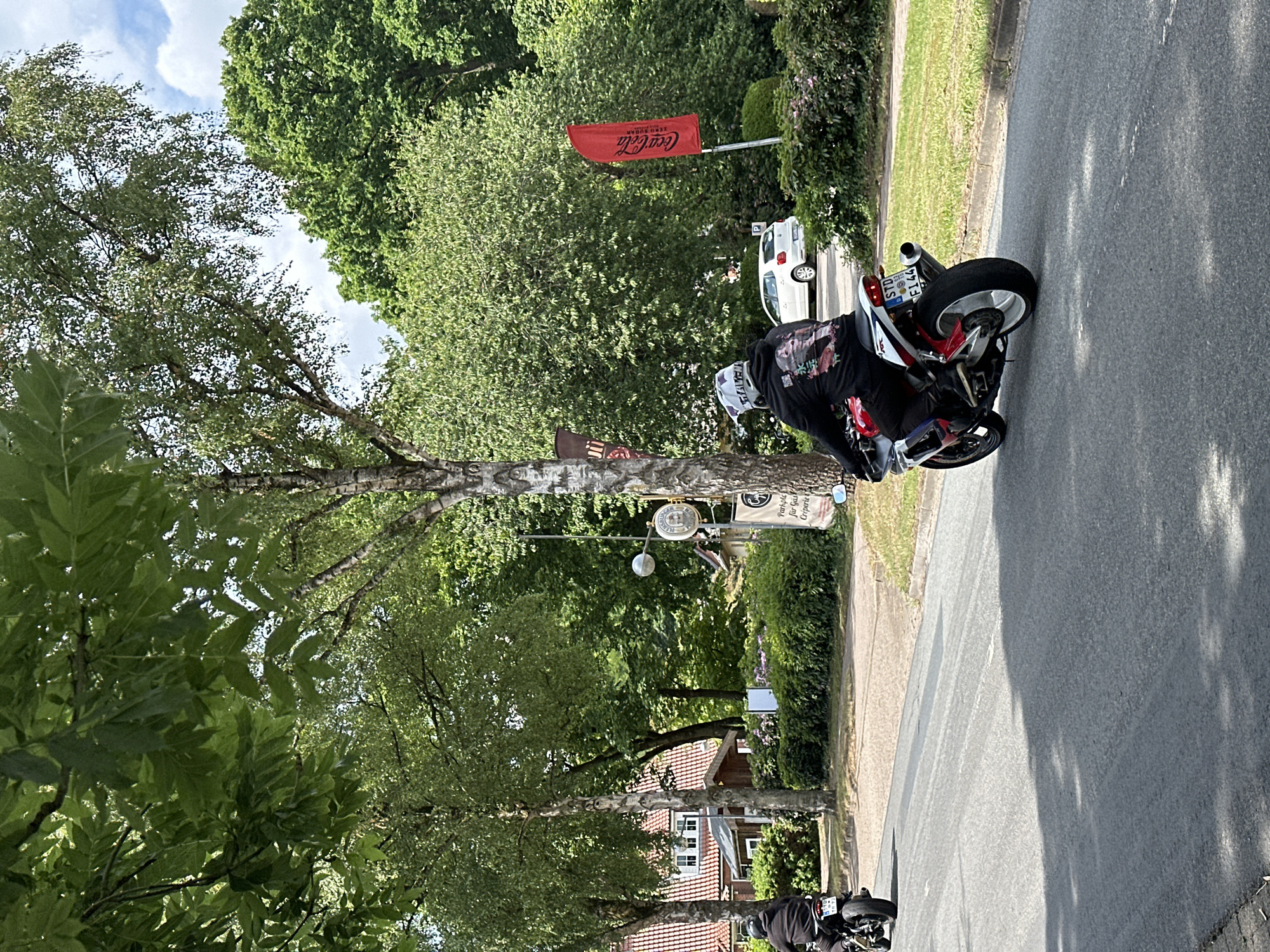 Two individuals on motorcycles are observed from behind, traveling away from the viewer on a paved road in Oberndorf, Germany. The scene depicts a tree-lined street under a partly cloudy sky.
The lead motorcyclist, positioned further down the road on the left, rides a dark-colored motorcycle. This rider wears a dark helmet and a dark jacket or hoodie. The motorcycle displays a German license plate reading "STD M 10".
The second motorcyclist, closer to the foreground and slightly to the right, operates a red and white sportbike with a white rear wheel and a silver exhaust pipe. This rider is wearing a white motocross-style helmet with a visible "FOX" brand logo on the back, and a black hoodie featuring a multi-colored graphic print of what appears to be an anime-style female character on the back. The motorcycle bears a German license plate stating "STD OS FI 44".
The road is asphalt, appearing dry with some visible tire marks and shadows cast by unseen objects to the left. A dirt and grass verge separates the road from the surrounding environment on the right.
To the right of the road, various signs and structures are visible. A red flag with the white "Coca-Cola" logo and the German text "WERBUNG / ERFRISCHT" is mounted on a pole. Further along, a small round sign resembling a traditional German inn or brewery emblem is affixed to a pole, below which is a black rectangular sign with white text advertising "Parkplatz für Gäste" (Parking for Guests) and "Crêperie". A white "P" (Parking) sign is also visible behind the second rider.
The background consists of lush green deciduous trees and bushes. Several houses with red tiled roofs and white facades are partially visible through the trees on the left side of the road. A white compact car is parked further back on the right, partially obscured by foliage. The overall environment suggests a suburban or semi-rural setting with established greenery.