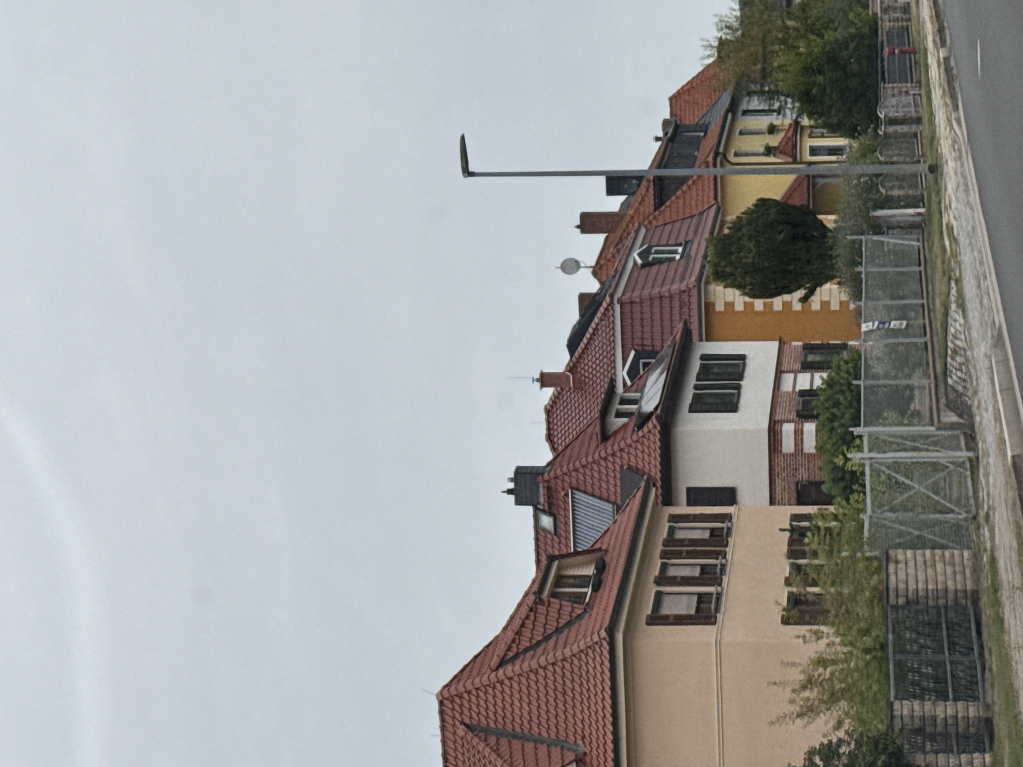 The image depicts a row of residential houses with red tiled roofs, characteristic of German architecture, located in Nordhausen, Germany. The houses vary in color and design, with some featuring stucco exteriors in light yellow and cream tones, and others with white facades and brick accents. Dormer windows and chimneys are visible on the sloped roofs. A modern street lamp stands to the right of the frame. The foreground shows a paved road and sidewalk, separated from the properties by a low brick wall topped with a metal fence and some greenery. The sky is overcast, suggesting a cloudy day. There are no people visible in the scene.