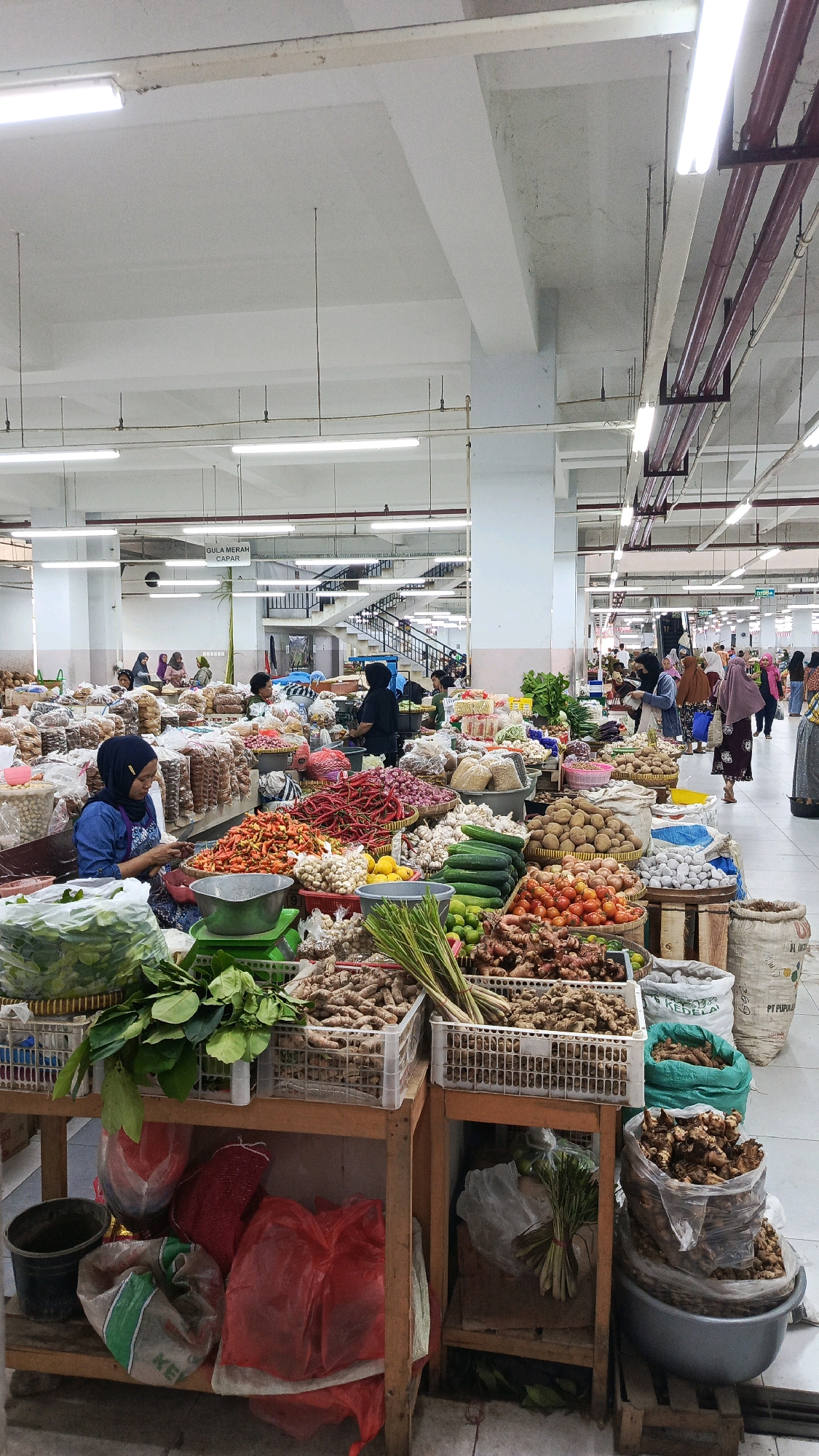 This is a full-shot, vertical-orientation photograph taken inside a covered market. The market is well-lit with fluorescent lighting fixtures mounted on the ceiling, which also has exposed pipes and beams. The market is filled with stalls displaying a wide variety of fresh produce. In the foreground, a woman wearing a dark blue shirt and a blue hijab is seated behind a stall laden with chilies, garlic, and lemons. To her left, a large basket overflows with green leafy vegetables. Behind her stall, other vendors are visible, as are customers browsing the merchandise.
The stalls are organized with produce displayed in baskets, crates, and large bags. Prominently featured are piles of red chilies, onions, potatoes, tomatoes, cucumbers, and various root vegetables like ginger and turmeric. Large sacks are also visible, some filled with produce and others appearing to contain grains or other dry goods. In the background, a set of stairs leads to an upper level of the market. People are seen walking, standing, and interacting with vendors, indicating a bustling marketplace environment. The overall impression is that of a lively and well-stocked local market, likely in Ponorogo, Indonesia, given the context.