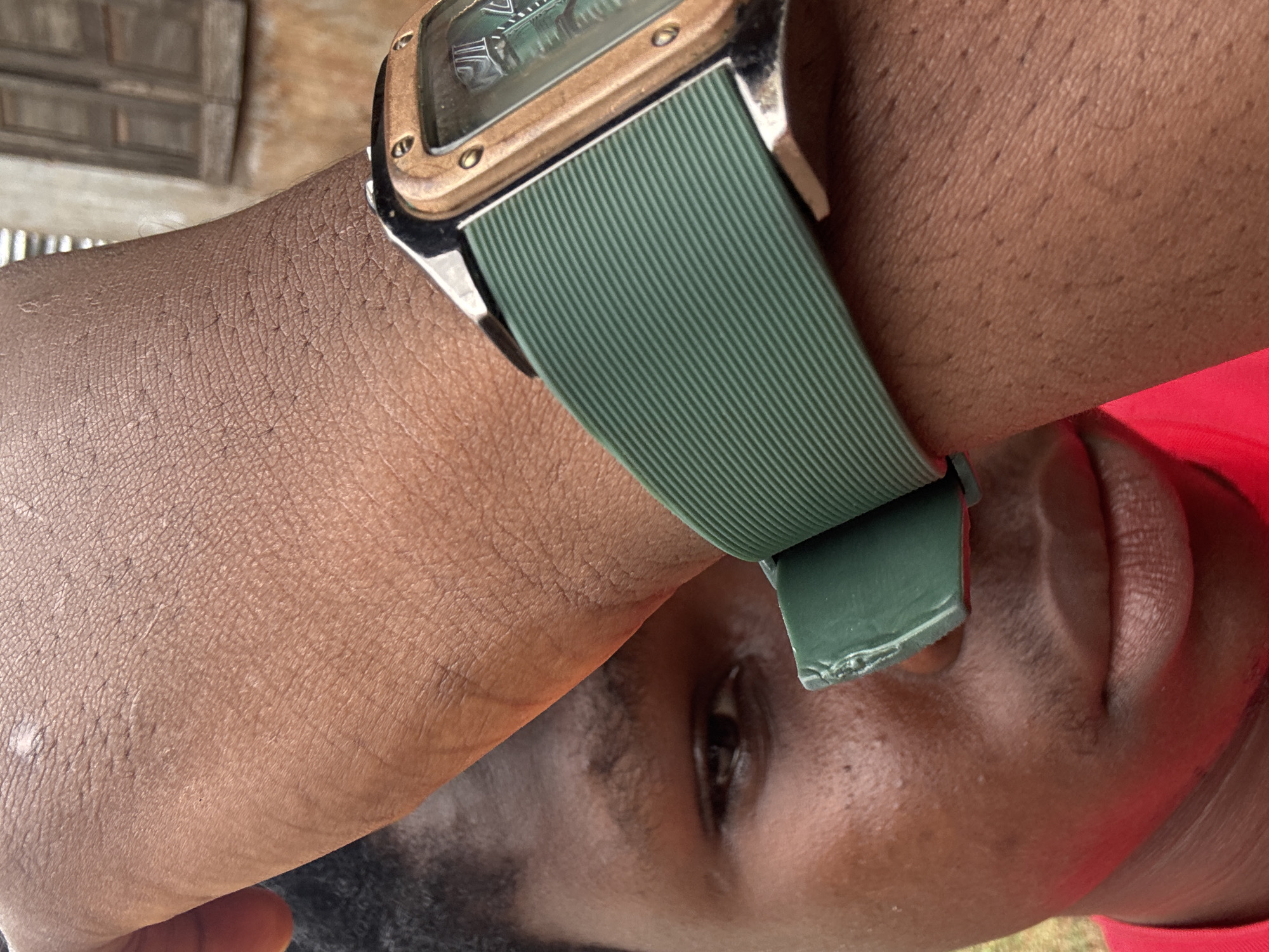 A close-up, head-and-shoulders shot captures a person wearing a green-strapped watch. The individual's face is partially obscured by their raised arm, revealing an eye and lips. The background shows a building with wooden shutters, possibly in Enugu, Nigeria. The lighting is natural, casting soft shadows.