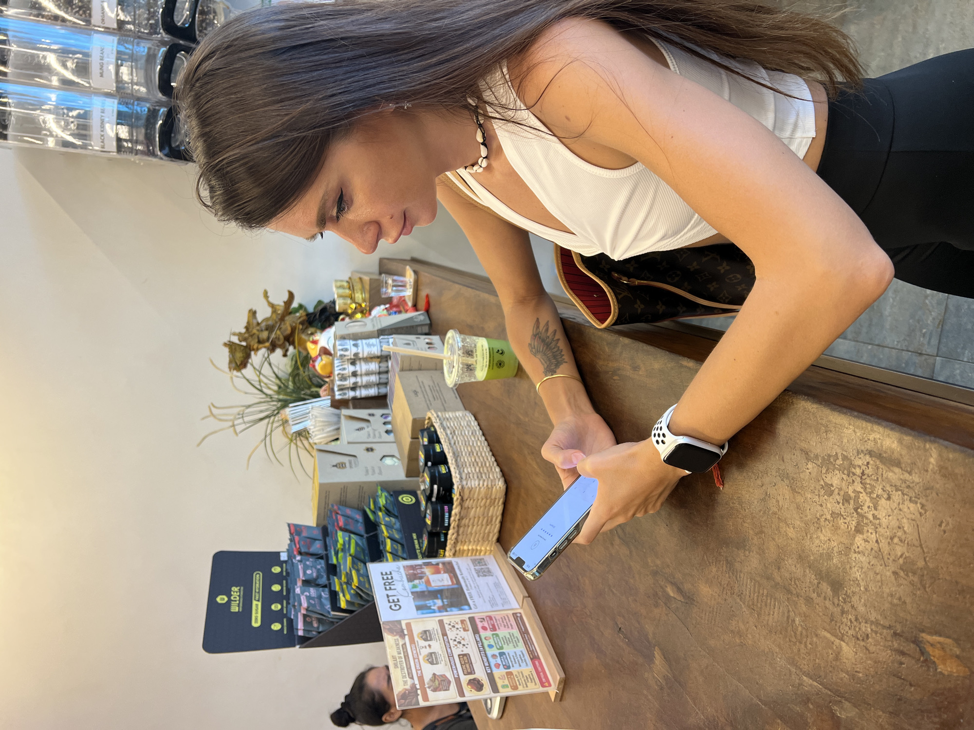 In a bright, casual setting, a young woman with long brown hair is looking down at her smartphone. She's wearing a white tank top and black leggings, with a white smartwatch on her left wrist. Her arm is resting on a wooden counter laden with various products in jars and boxes. Some of the products have labels that appear to be in English. A woven basket filled with small jars sits on the counter, and a green beverage in a plastic cup with a straw is also visible. A magazine or brochure is open on the counter in front of her, displaying colorful graphics and text. In the background, shelves stocked with more items can be seen. The overall scene suggests a retail environment, possibly a health food store or a market, in the Ko Samui District of Thailand. The lighting is natural and suggests daytime. The woman appears focused on her phone, with a neutral expression.