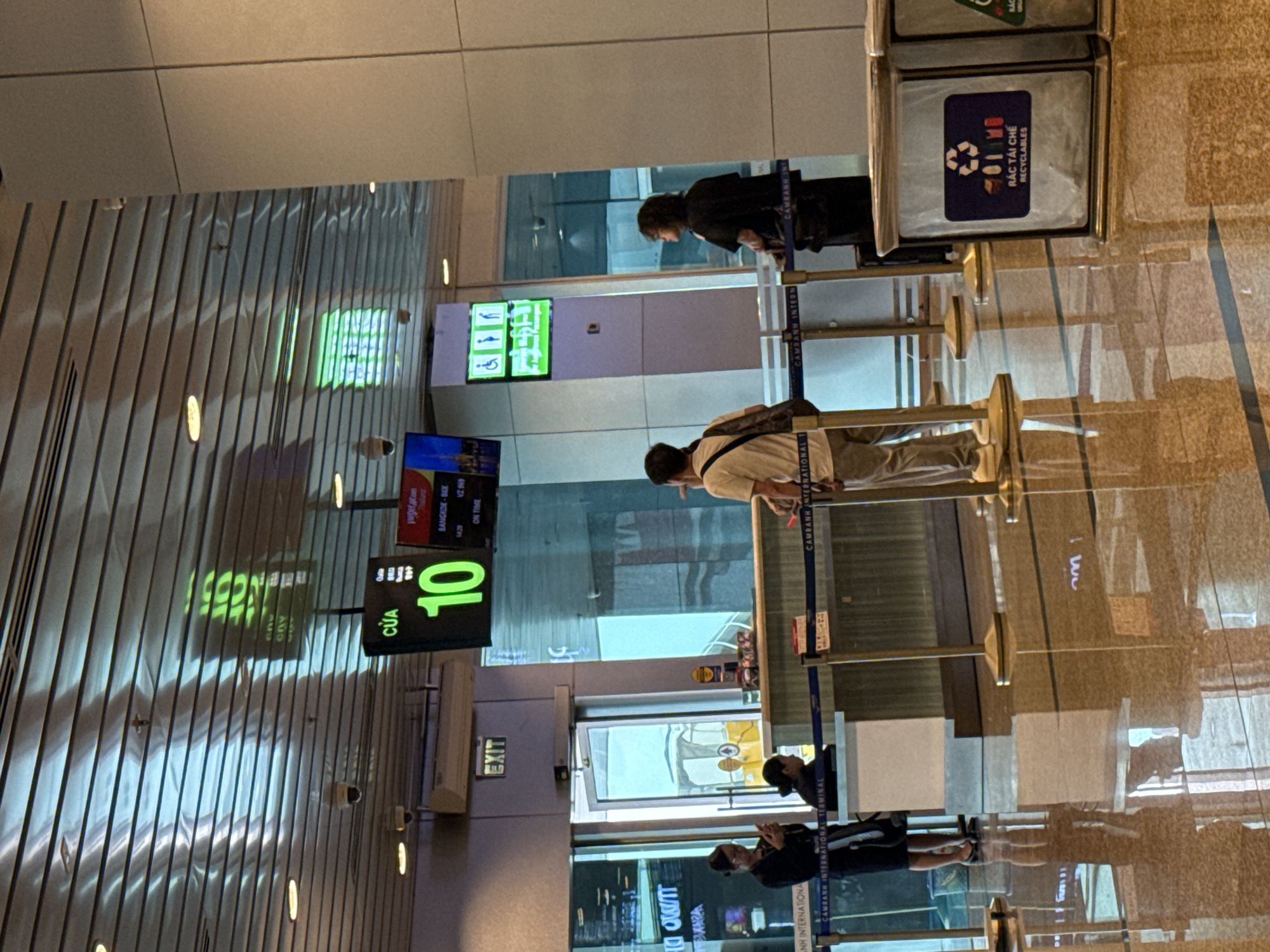 The image shows a bright, modern airport terminal interior. The ceiling features a distinctive ribbed metal design with embedded lighting. In the background, a digital display board shows flight information, with "CUA 10" (Gate 10) prominently displayed in large green numerals. To the right of this display, a smaller screen shows "VegeCar.com" with "BANGKOK - BKK" and "ON TIME" details, suggesting flight information. Further to the right, a green sign displays accessibility symbols for people with disabilities.
In the mid-ground, a counter area is visible, with several people interacting. Two women are behind the counter, one of whom is looking at her phone. A man with a backpack is standing in front of the counter, facing away from the camera. Another person, a woman with dark hair, is standing to the right of the counter, also facing away.
The floor is highly polished and reflective, showing the reflections of the people and objects in the terminal. To the right of the scene, two large recycling bins are visible. One bin has a blue label with the recycling symbol and the text "RÁC TÁI CHẾ RECYCLABLES" in Vietnamese and English.
The overall impression is of a busy, functional airport environment. The presence of Vietnamese text and the "CAM RANH INTERNATIONAL TERMINAL" markings on the stanchions strongly indicate the location as Cam Ranh International Airport, located in Cam Ranh city, Khánh Hòa Province, Vietnam.