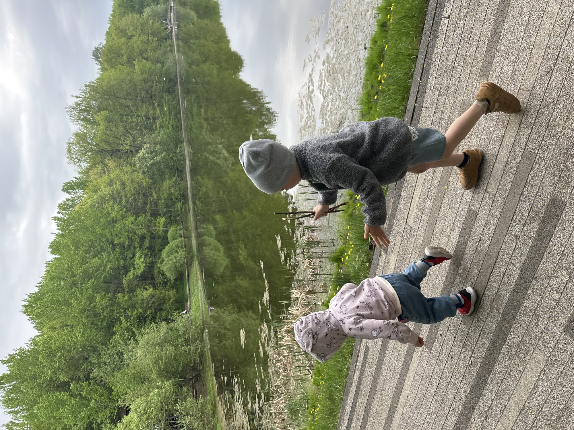Two children are near a body of water in Nizhny Novgorod, Russia. One child, wearing a gray beanie and a gray fleece, is holding sticks. The other child, wearing a light pink hoodie with a rabbit pattern and blue jeans, is lying prone on the paved path. The body of water is partially covered in vegetation. Trees line the opposite bank of the water.