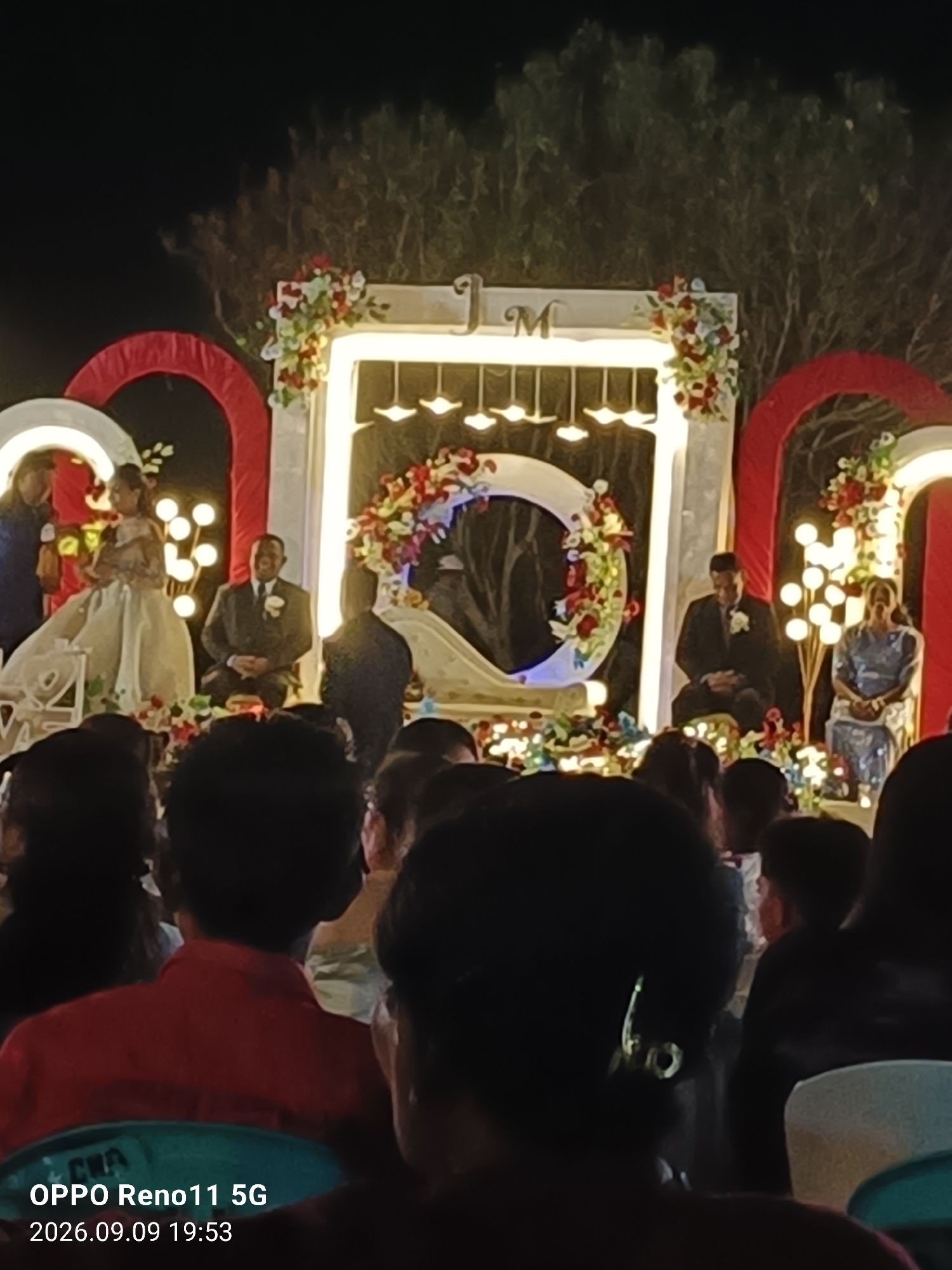 An outdoor evening wedding reception is captured from a low angle, looking towards a stage decorated with floral arrangements and large white arches. In the center of the stage, a bride in a white gown stands next to a groom in a dark suit. Behind them, a large white crescent moon decoration is visible. To the left of the bride, a person in a dark suit stands, possibly an officiant. To the right of the groom, a woman in a blue patterned dress is seated. The stage is illuminated by string lights and spotlights. In the foreground, the backs of several guests are visible, seated in chairs. The overall atmosphere appears to be festive, with a focus on the wedding ceremony. The image contains text overlay in the bottom left corner indicating "OPPO Reno11 5G 2026.09.09 19:53". The location is identified as Otan, Indonesia.