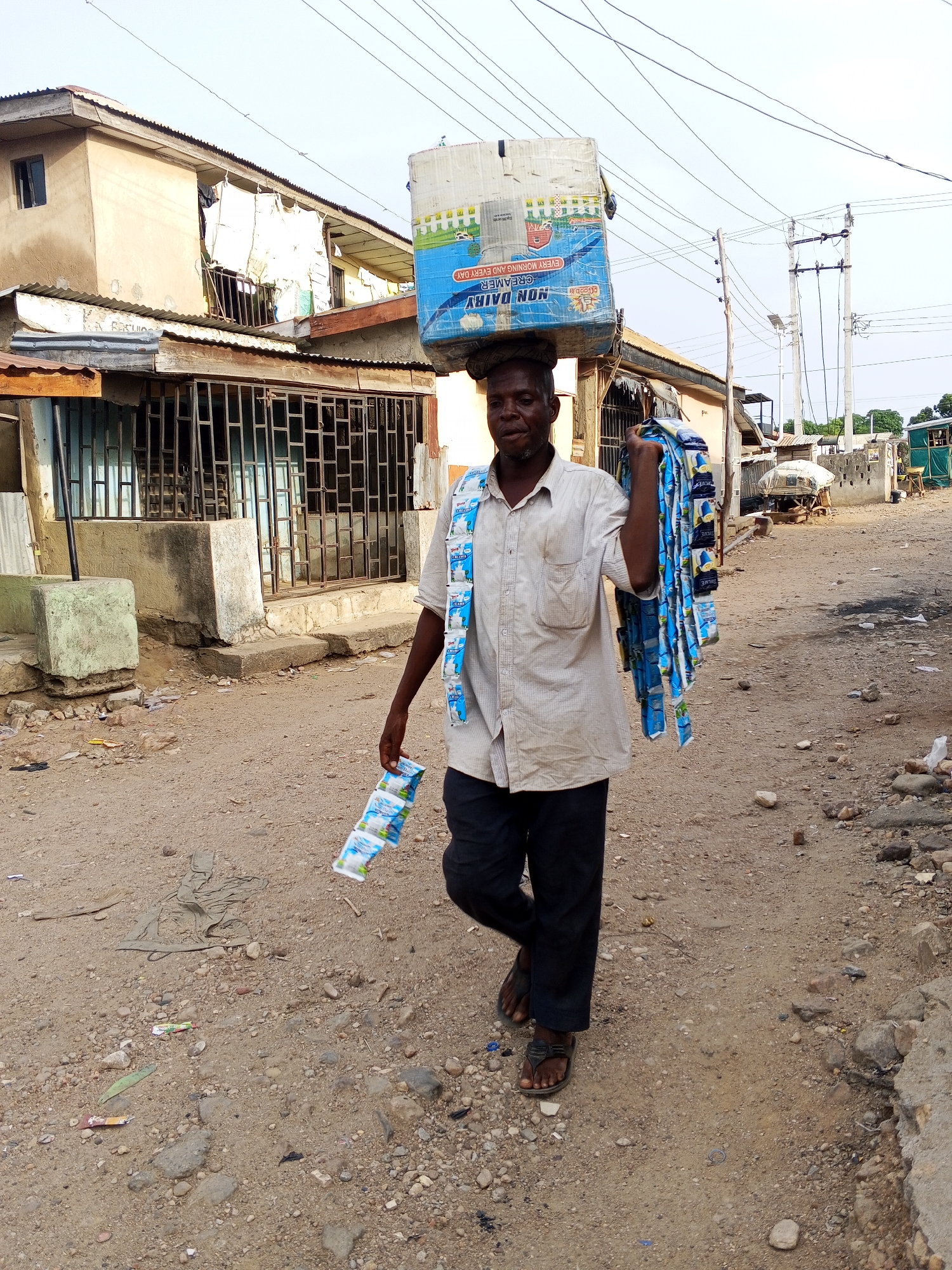 An adult male walks along a dirt road in Adankolo, Nigeria. He carries a large box labeled "NON DAIRY CREAMER" on his head and numerous individual packets of what appear to be the same product hanging from his body. The street is lined with simple, weathered buildings. Trash and debris litter the ground. Overhead, power lines are visible. The lighting suggests it is daytime.