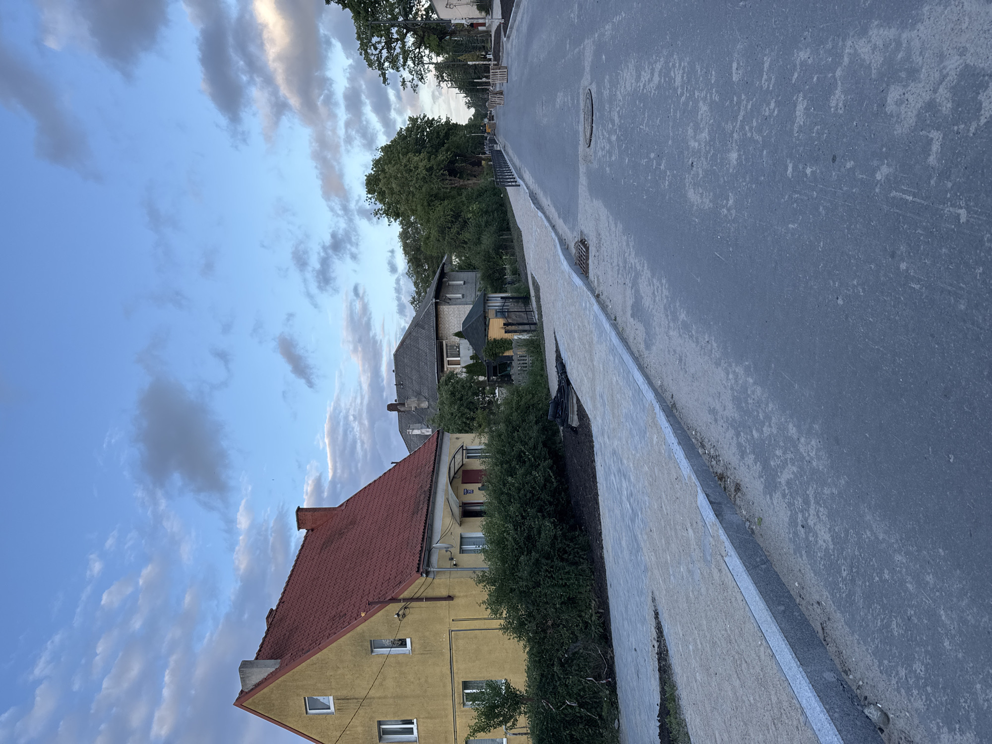 The image depicts a street scene in Guryevsk, Russia, during what appears to be late afternoon or early evening. The sky is partially cloudy with a mix of blue and grey.
In the foreground, a new asphalt road is visible, with a newly constructed sidewalk or pedestrian path on the left. The sidewalk is made of light-colored paving stones and bordered by a granite curb. Adjacent to the sidewalk, there is a strip of soil and some landscaping materials.
Several houses are visible along the street. On the left, a two-story yellow house with a red tiled roof and white-framed windows stands out. Further down the street, other houses with pitched roofs can be seen. Some trees and dense green bushes line the property borders.
The road itself appears to be undergoing some form of construction or renovation. There are manhole covers visible in the asphalt. In the distance, there are some construction materials, including pallets and possibly yellow construction equipment. Streetlights and utility poles are present along the side of the road. The overall impression is of a residential area in transition, with infrastructure improvements underway. There are no people visible in the image.