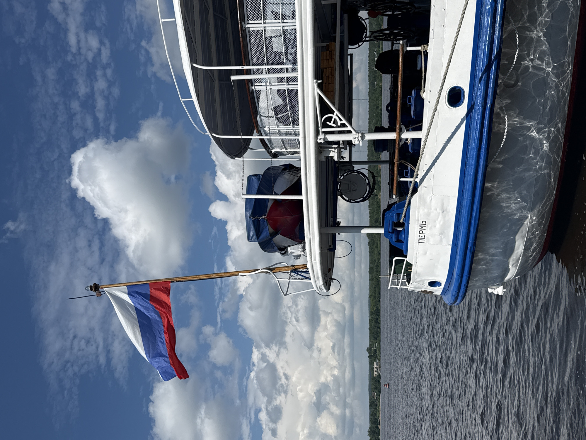An outdoor, low-angle, medium shot shows the front of a white and blue pleasure boat docked on dark, rippling water under a bright blue sky with scattered white clouds. A Russian flag flies from a pole at the front of the boat. The boat has multiple decks with white railings and a white awning. A small, red and blue covered inflatable boat is strapped to the top deck. The name "ПЕРМЬ" is visible in black letters on the white hull of the boat. In the background, a green, tree-lined shore with buildings is visible across the water, with a red and white buoy in the foreground. The sun glints off the water, creating dappled light on the lower hull of the boat. There are no people visible in the frame.