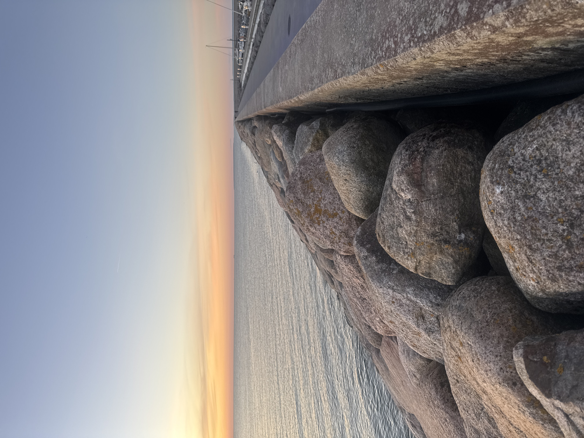 This is a photograph of a stone breakwater extending into the sea during a sunset. The sky is a gradient of pale blue to orange. The water is calm and reflects the colors of the sky. The breakwater is made of large, rough-hewn stones. To the right of the breakwater is a paved walkway and a marina with several sailboats docked. There are a few indistinct figures walking on the walkway in the distance. The scene appears to be in Tallinn, Estonia, a city known for its coastal location and harbor. The photograph is taken from a low angle, emphasizing the scale of the breakwater and the expanse of the sea and sky.
