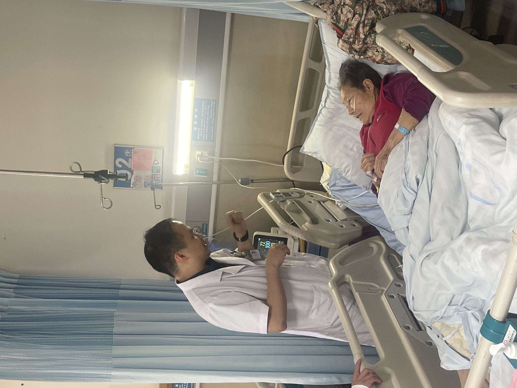 An elderly Asian woman is in a hospital bed. She has a nasal cannula in her nostrils and a blue wristband on her left wrist. She is lying under white bedding and wearing a red long-sleeved shirt. To her left is a doctor or medical professional in a white coat. He is facing her and gesturing with his right hand. A bedside monitor displays patient vitals. The hospital room has blue curtains, an IV pole, and a fluorescent light. There is a sign on the wall with Chinese characters. This scene is in a hospital room in Tongliang District, China.