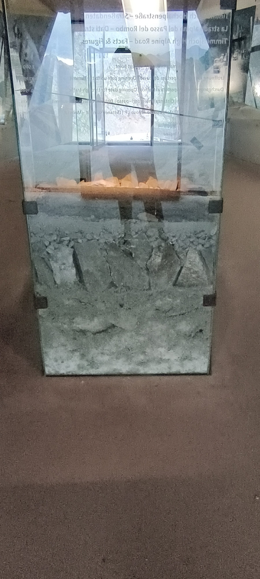 The image shows a glass display case in Sölden, Austria, exhibiting a cross-section of soil layers, likely a geological or historical exhibit. The display case is transparent, revealing layers of sediment and rocks. The top layer appears to be composed of light-colored stones, possibly gravel, set on a darker, possibly sandy or loamy soil layer. Beneath this, there are thicker layers of broken rock and compacted soil, with evidence of stratification. The exhibit is illuminated from within or above, casting shadows within the case. The background, visible through the display case, shows a building interior with glass doors and reflective surfaces, suggesting a museum or visitor center. The floor in front of the display case is a reddish-brown carpet. The text on the display case, though partially obscured and reversed, appears to be informational signage related to the exhibit.